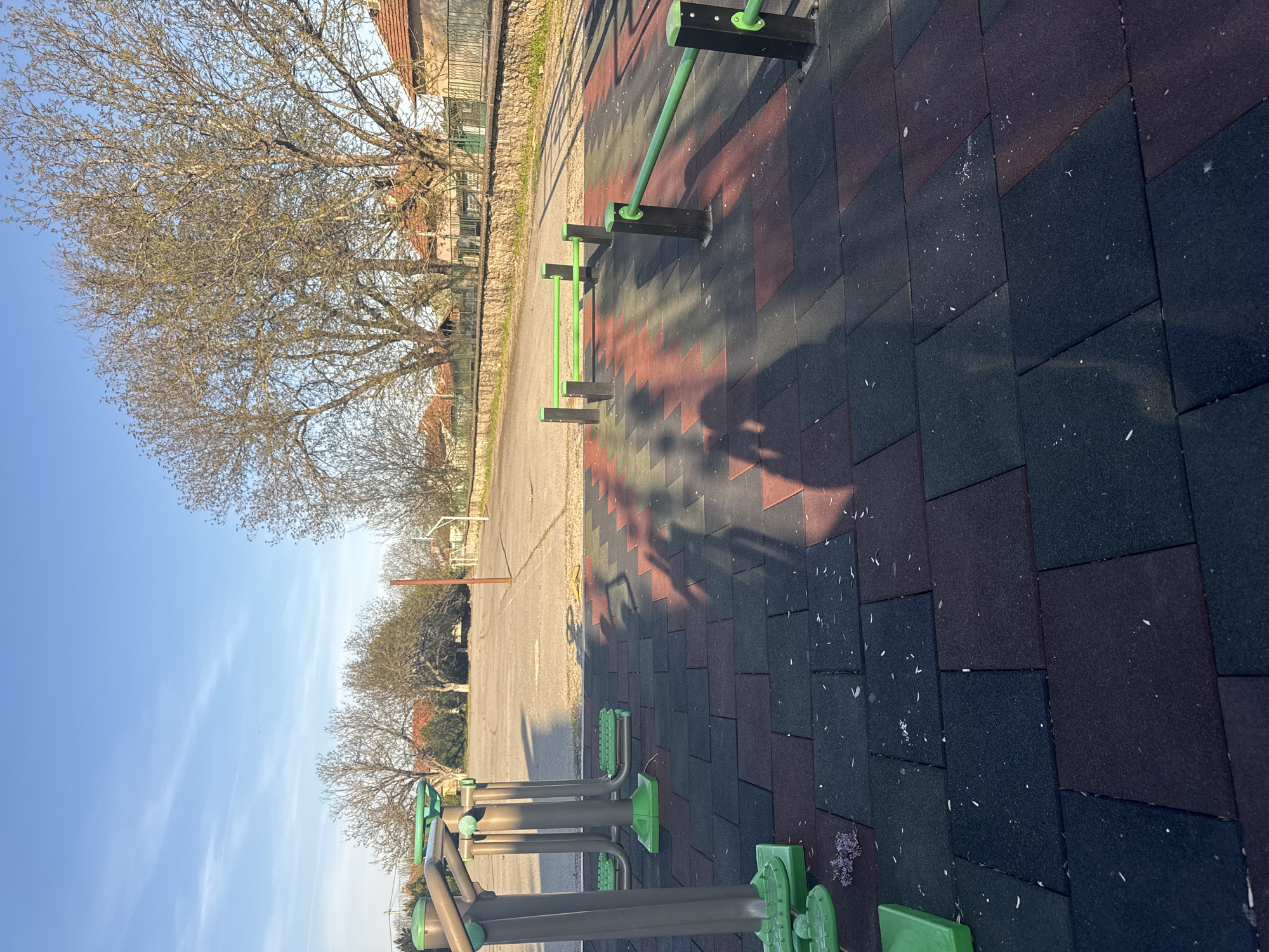The image shows a sunny outdoor workout area in Lehchevo, Bulgaria. The ground is covered with interlocking rubber tiles in shades of black and red. Exercise equipment with green accents is visible in the foreground and middle ground. A paved area extends beyond the workout space, with trees visible on either side. Buildings are visible in the distance behind the trees. The sky is clear blue.