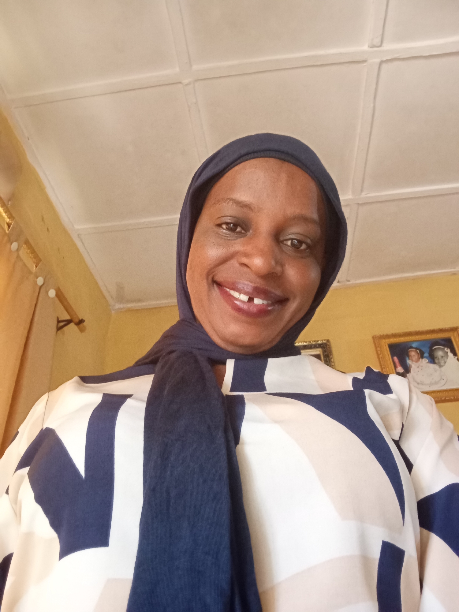A medium shot captures a smiling woman wearing a dark blue hijab and a patterned top with navy blue and off-white geometric shapes. She is positioned indoors, likely in a home in Narayi, Nigeria, judging by the background elements. Behind her, a yellow wall is visible, adorned with framed pictures. The ceiling is white with recessed square panels. A curtain with a wooden rod hangs slightly to the left.