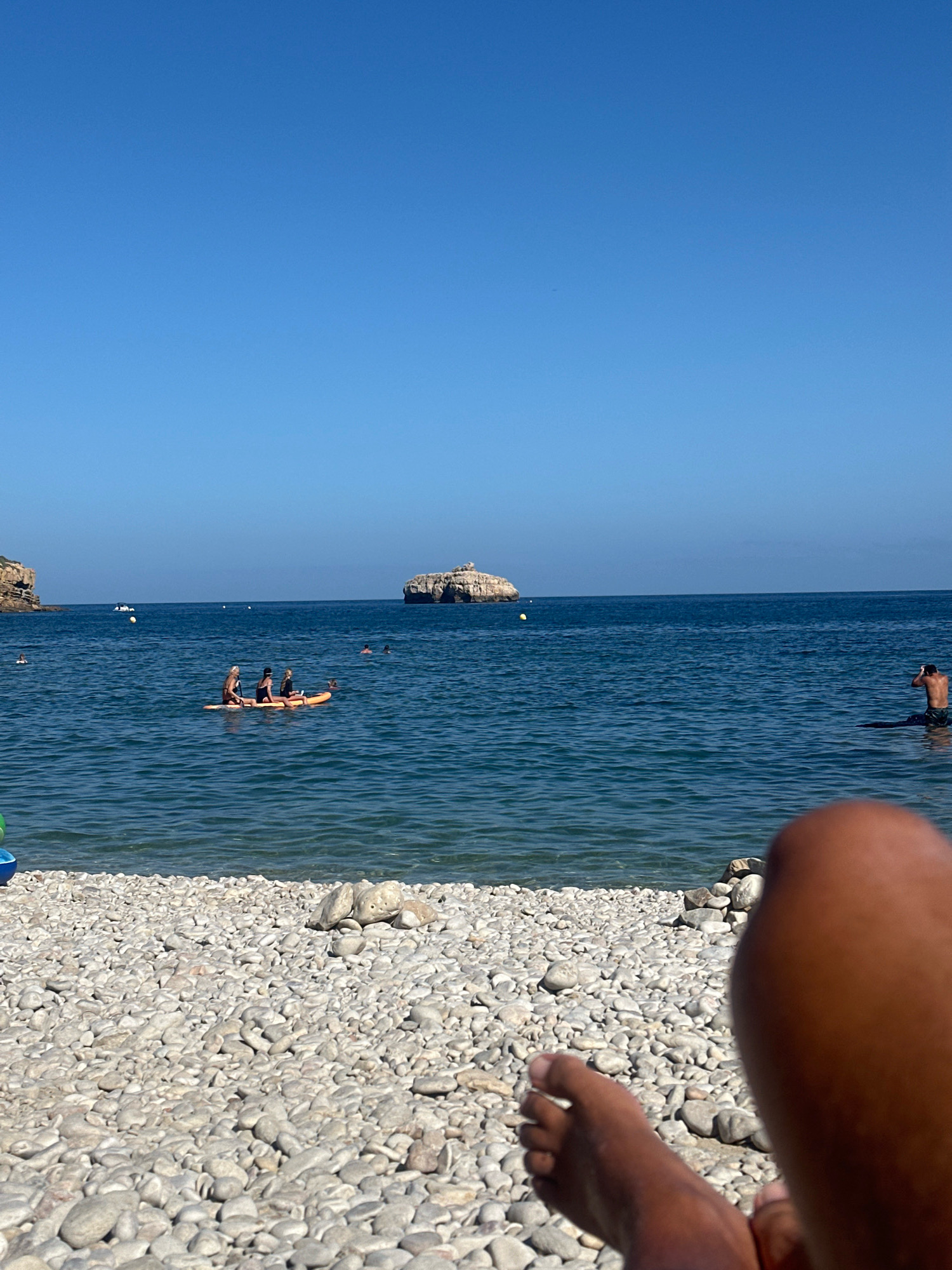 The image depicts a sunny day at a pebbled beach in Xàbia / Jávea, Spain. In the foreground, a person's legs and feet are visible, suggesting they are relaxing on the beach. The mid-ground shows a calm, blue sea with several people engaged in water activities. A yellow inflatable raft with three individuals is in the center, and another person is standing in the water to the right. In the background, a rocky island formation stands in the sea, with coastal cliffs visible on either side of the bay. The sky is clear and bright blue.