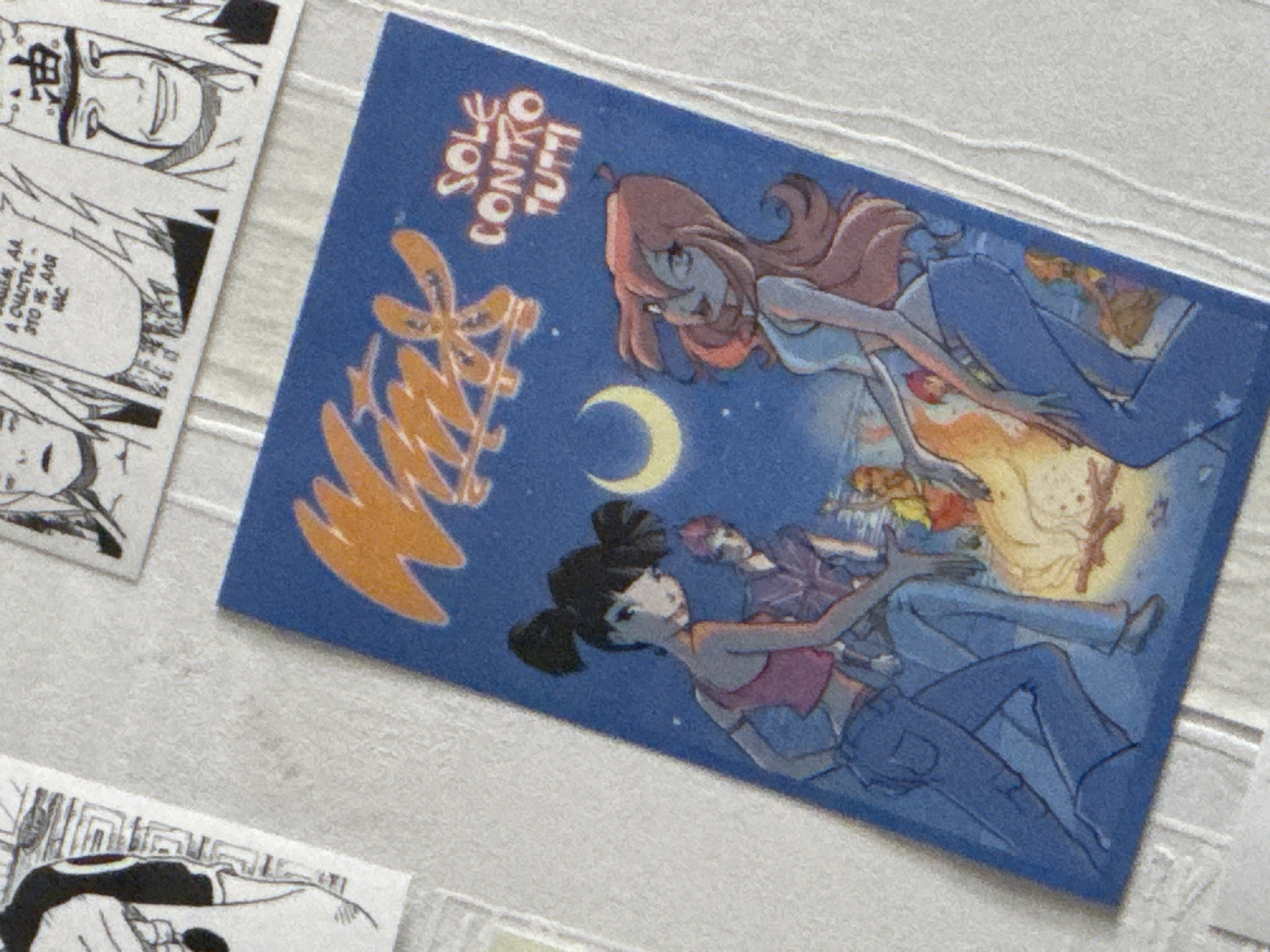 The image is a close-up of a collection of stickers or trading cards arranged on a textured white surface. The main subject is a blue card featuring an illustration of four young women and a bonfire under a crescent moon. Two of the women are prominently featured in the foreground, dressed in denim outfits and sitting near the fire. One has black hair in buns and wears a pink top, while the other has long, reddish-pink hair and a light blue top. In the background, two other figures are partially visible near the fire, and the text "SOLE CONTRO TUTTI" is written in white stylized lettering on the right side of the card, above the women. The overall style of the illustration appears to be anime-inspired. Other cards or images, some of which appear to be black and white manga panels, are visible in the upper left and upper right portions of the frame, partially overlapping the central blue card. The lighting is diffuse and appears to be natural. The context of Moscow, Russia, is not present in the image itself and cannot be inferred from the visual information.