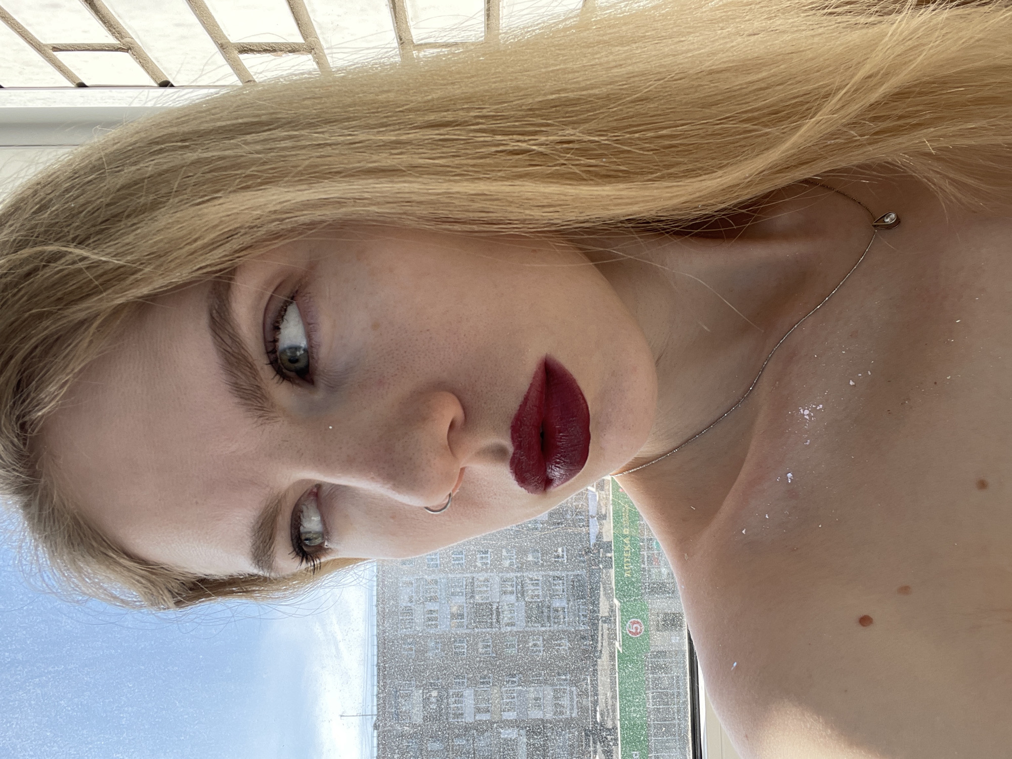 A close-up indoor portrait features a female-presenting individual positioned against a window overlooking an urban landscape. The individual has long, straight blonde hair, a fair complexion, and blue-green eyes directed towards the left side of the frame. A silver-colored hoop nose ring is present in the left nostril, and the lips are colored with dark, matte burgundy lipstick. Scattered reflective particles, possibly glitter, are visible on the right cheekbone and the exposed right shoulder and collarbone. Several brown moles are present on the right shoulder and upper chest. A thin, silver-colored necklace with a small, teardrop-shaped pendant containing a clear stone is worn around the neck.
The background, viewed through a partially obscured window, reveals an urban environment under a clear blue sky. A large, grey, multi-story building with numerous windows occupies the left side of the background. At street level, a commercial establishment features a prominent green sign with Cyrillic text, clearly displaying "АПТЕКА" (Pharmacy) and a red circle containing the number "5," indicative of a typical urban scene in Izhevsk, Russia. A white tiled wall or structure is visible on the far right of the frame, adjacent to the individual.