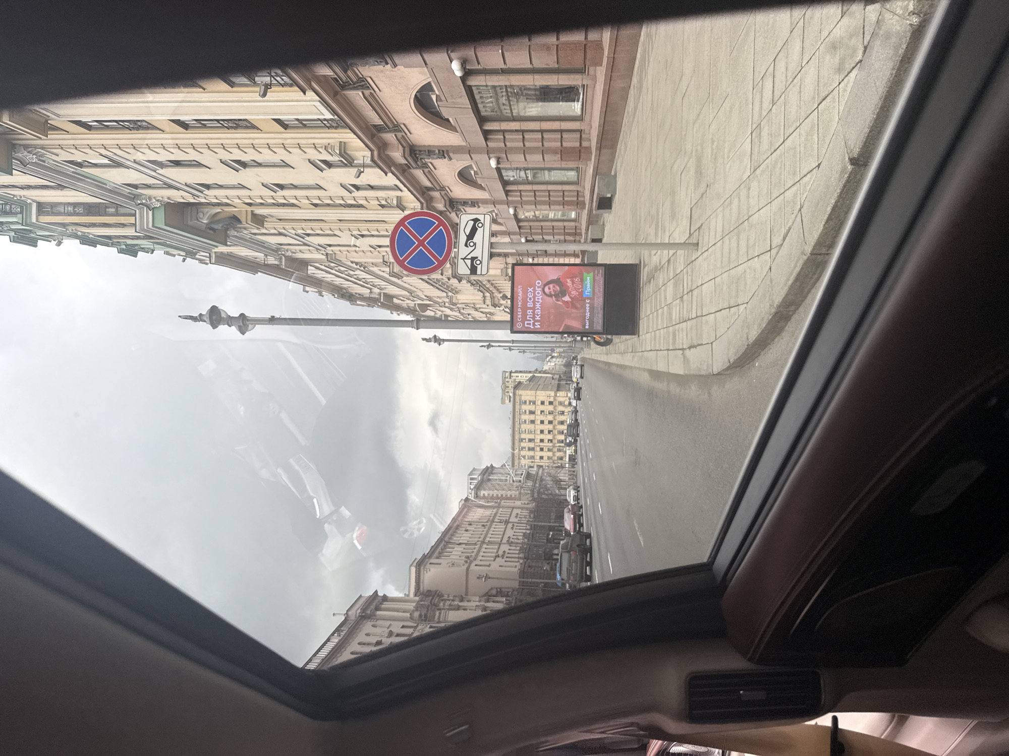 This media file captures a street scene in an urban environment, viewed from inside a vehicle. The image appears to be taken looking out from a sunroof or a high window, offering a perspective of buildings, a street, and an overcast sky. The dark frame of the car's interior is visible at the top and bottom, along with an air vent in the lower right, and reflections on the glass.
The street is bordered by grand, multi-story buildings exhibiting classical European architecture, characterized by ornate facades, numerous windows, and decorative cornices, predominantly in light beige and cream tones. A wide sidewalk, paved with light square tiles, runs along the base of these structures.
Prominently featured on the street are several signs. A circular "No Stopping" sign (a red circle with a blue cross) is clearly visible, accompanied by a smaller sign depicting a tow truck, indicating that stopped vehicles will be towed. Adjacent to these signs stands a vertical digital billboard displaying a smiling woman. The billboard features Cyrillic text, reading "СБЕР МОБАЙЛ" (SberMobile), "Для всех и каждого" (For everyone), and "ВЫГОДНЕЕ С ПРИЯНО" (More profitable with Priyano or pleasantly).
Further down the road, more buildings and several vehicles, both parked and in motion, can be seen, contributing to the sense of a busy city street. The sky is uniformly overcast, suggesting a cloudy daytime, though the exact time is not discernible. The combination of the distinctive architecture and the Cyrillic text on the billboard confirms the setting as Moscow, Russia.