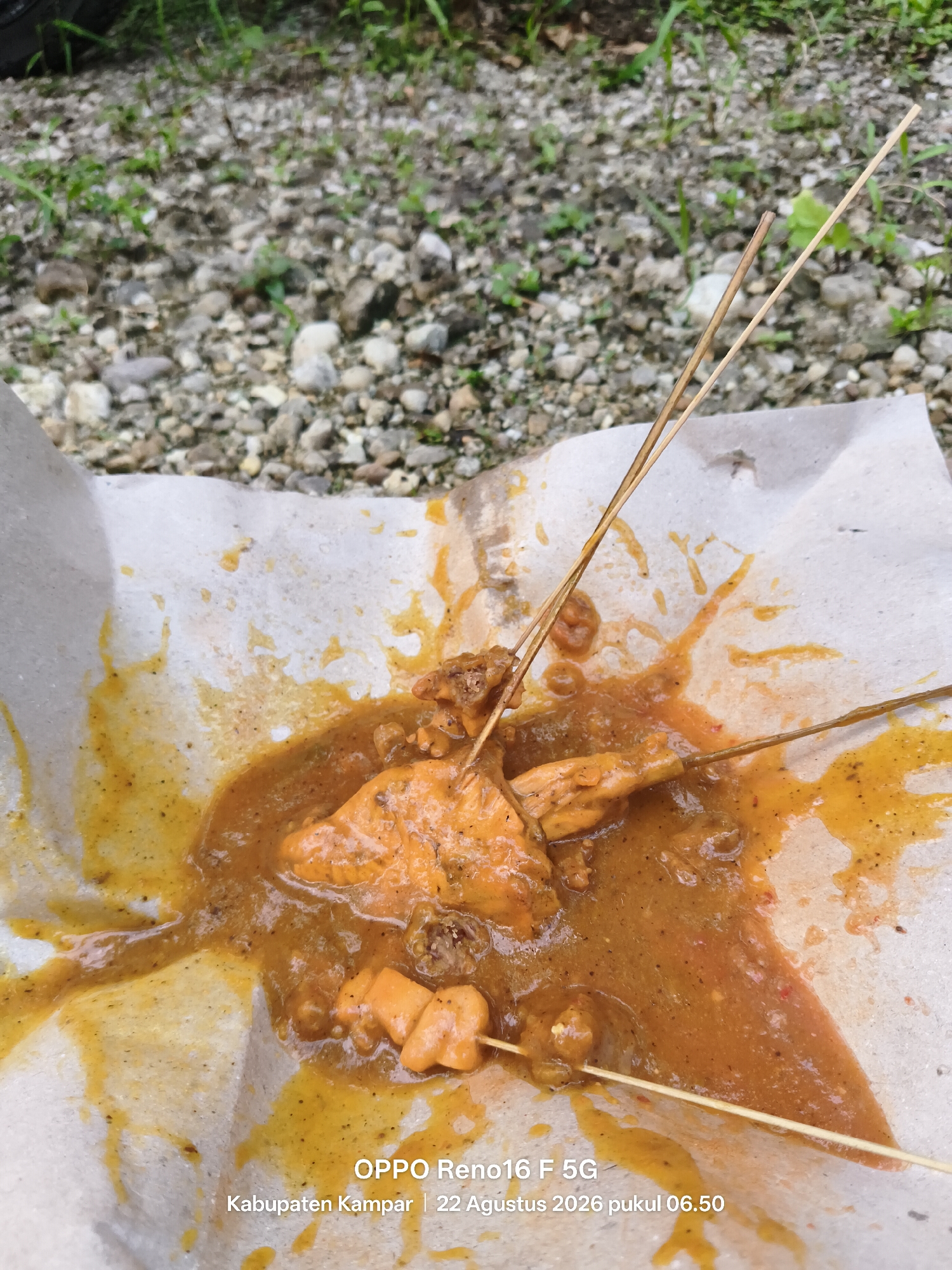 A close-up, overhead view of a plate of chicken satay, generously coated in a thick, orange-brown peanut sauce, is presented on a piece of textured paper. The satay skewers, made of thin, light-brown wood, pierce through chunks of chicken, some of which appear to be skin and others more solid meat. The sauce has spilled and spread across the paper, with visible streaks and pools. Small, dark specks, possibly ground spices or pepper, are dispersed throughout the sauce. The background is blurred, showing a rough, gravelly surface with scattered small stones and green vegetation, suggesting an outdoor setting. Text overlays the bottom of the image, indicating "OPPO Reno16 F 5G," and the location "Kabupaten Kampar | 22 Agustus 2026 pukul 06.50," which translates to Kampar Regency | August 22, 2026 at 06:50 AM.