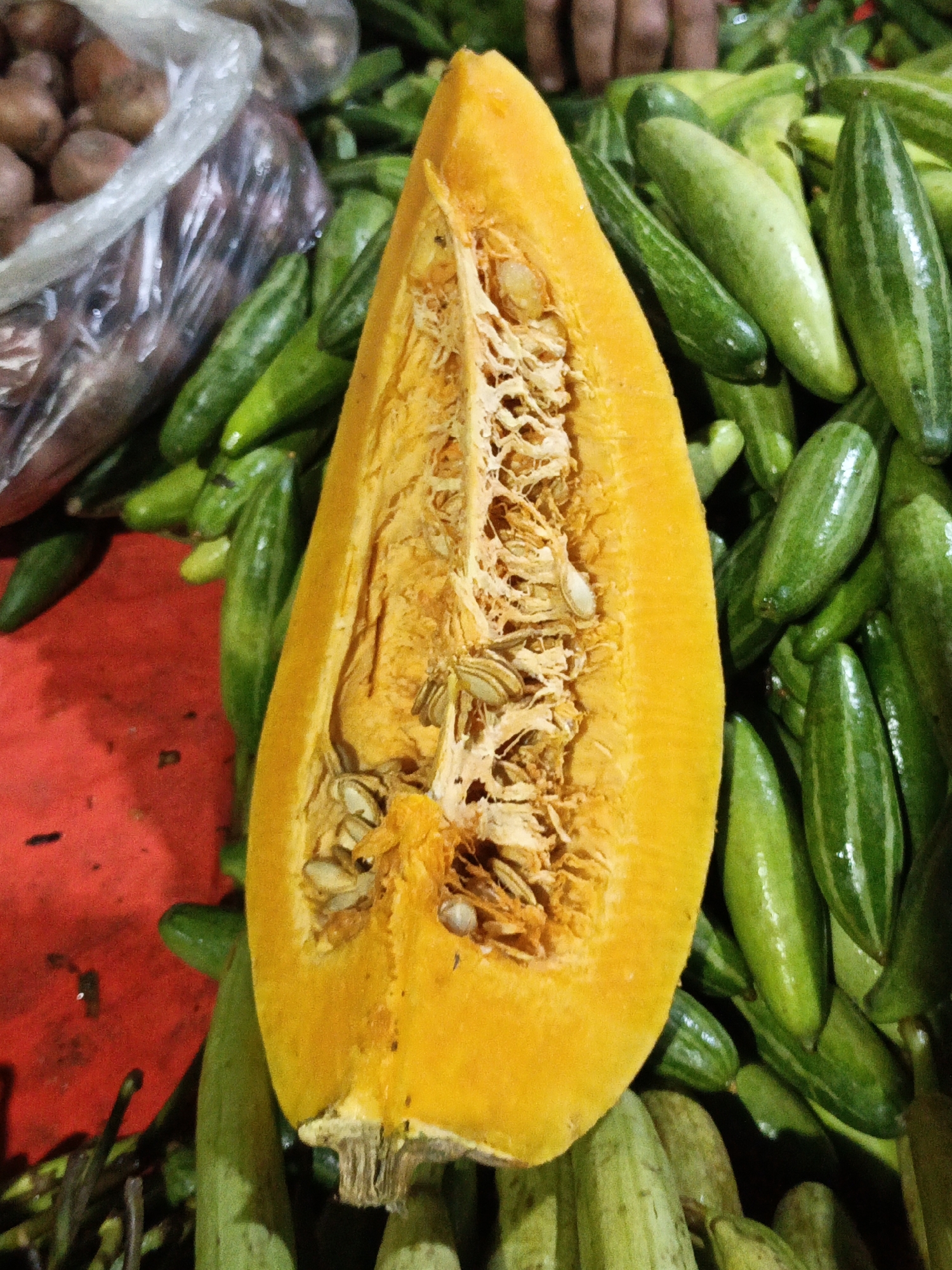 A close-up, top-down shot captures a vibrant market scene in Dhaka, Bangladesh. The focal point is a bright orange pumpkin, sliced in half, revealing its fibrous interior and numerous seeds. This prominent pumpkin rests atop a bed of numerous green, elongated vegetables, likely gourds or cucumbers, their smooth skins gleaming under the light.
To the left of the pumpkin, a plastic bag filled with small, reddish-brown onions is visible. The background is a blur of more green vegetables and hints of a reddish surface, possibly a market stall or tarp. The lighting suggests an indoor or shaded outdoor setting, typical of a bustling marketplace. A person's hands are partially visible in the upper background, suggesting ongoing activity, perhaps a vendor or shopper interacting with the produce.
The image conveys a sense of abundance and the everyday commerce of fresh produce in a South Asian market. There is no visible text in the image.