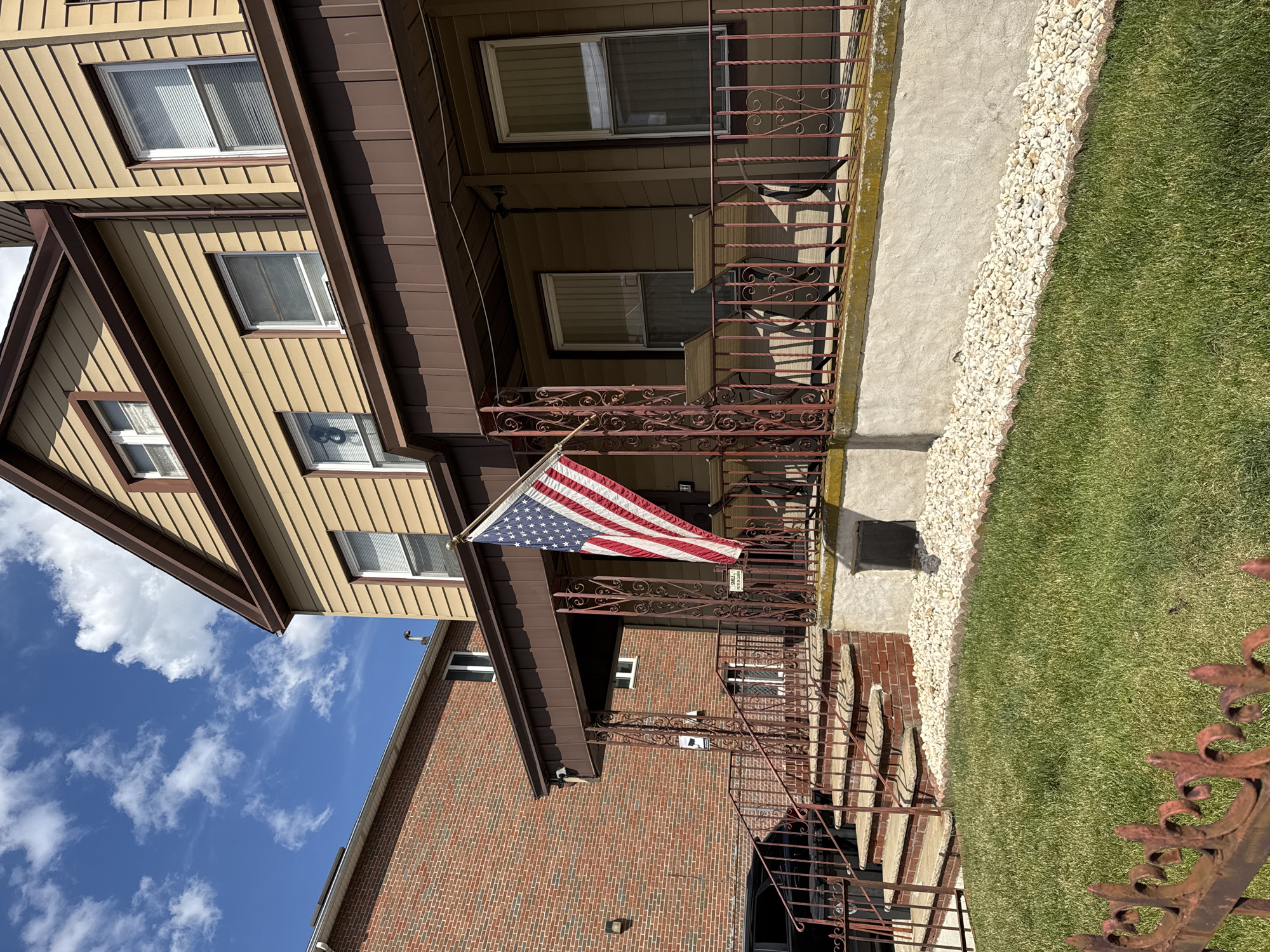 A two-story house with beige siding and brown trim stands under a partly cloudy blue sky. An American flag hangs from a pole on the front porch, which is enclosed by a brown metal railing with ornate scrollwork. The porch has two chairs and a small table. To the left of the house, a brick building is visible. A green lawn in front of the house is bordered by a decorative metal fence and a bed of white pebbles. Stairs lead up to the porch from the side yard. The image appears to be taken in a residential neighborhood.