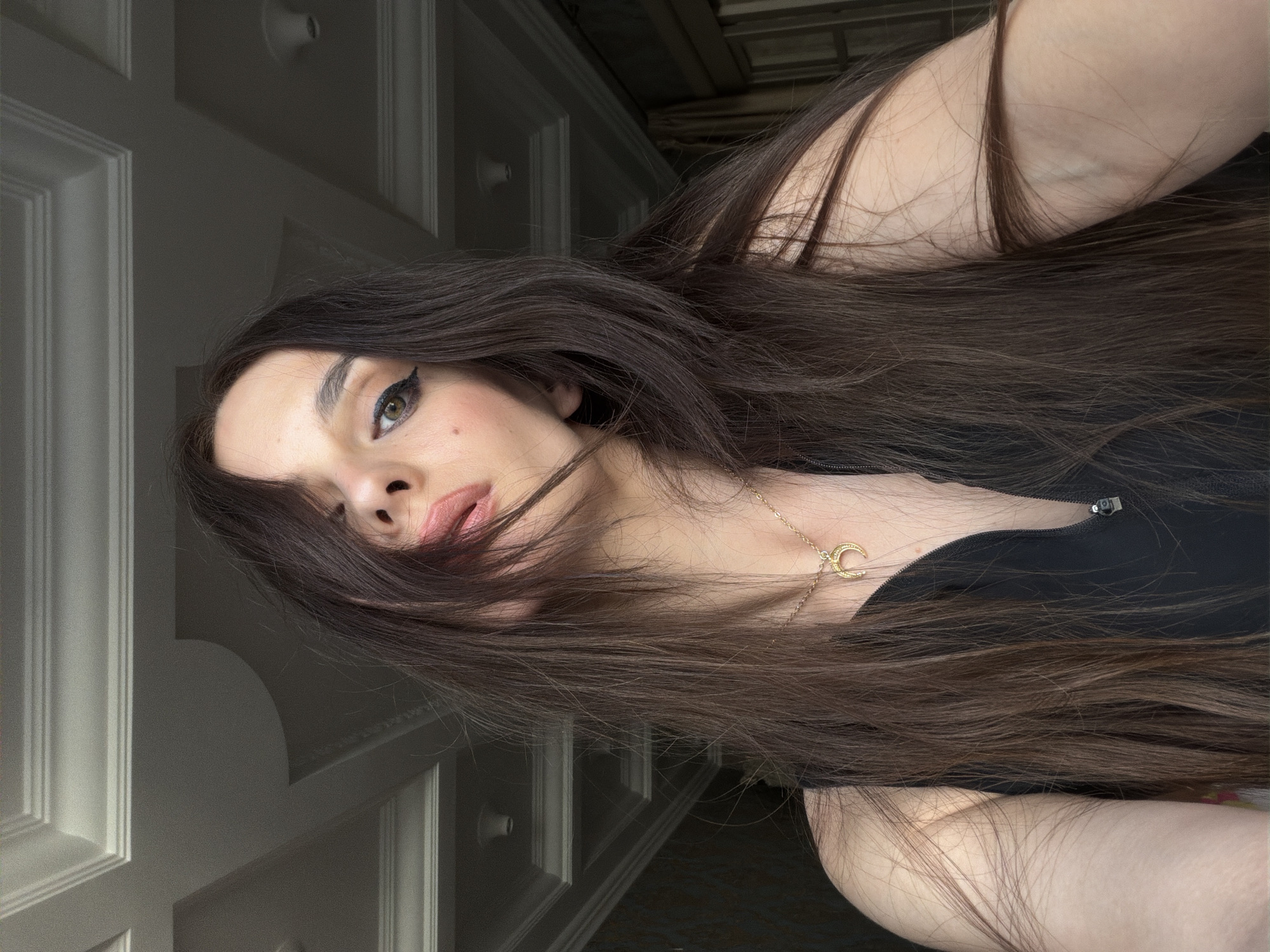 A woman with long, dark brown hair is shown from the chest up. She is wearing a black zip-up top and a gold crescent moon necklace. Her hair is partially covering her face. She is wearing eye makeup, including winged eyeliner. The background appears to be indoors, with decorative ceiling molding and patterned wallpaper visible. The lighting suggests it is daytime, possibly near a window. No specific location in Ufa, Russia, can be determined from the image.