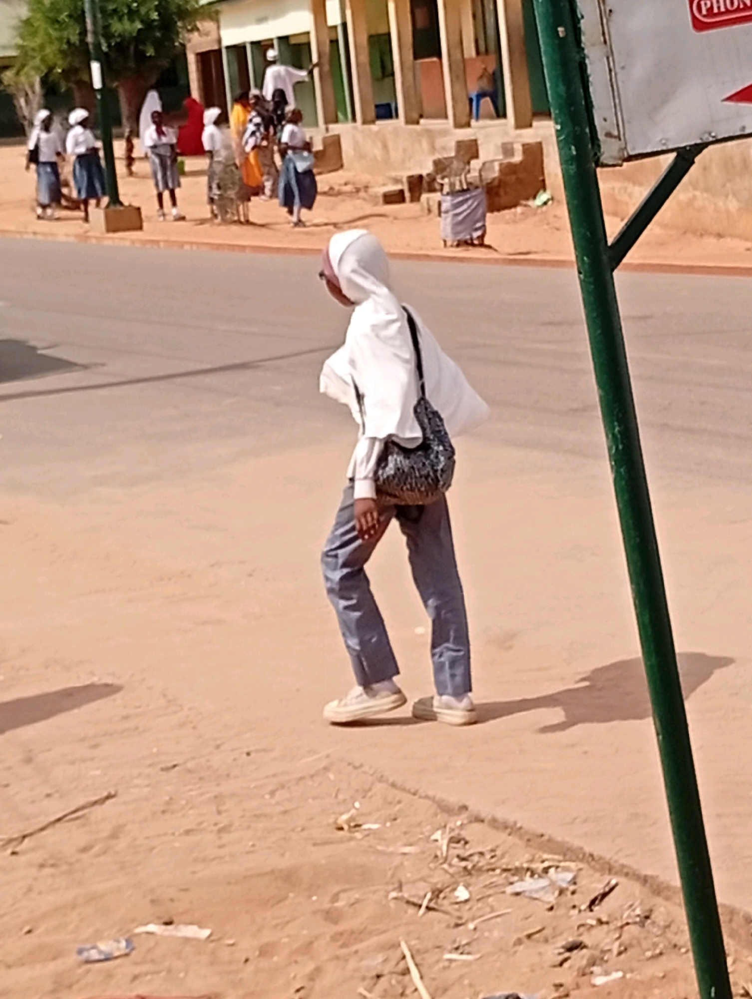 The image depicts a street scene in Gombe, Nigeria. A person wearing a white hijab and a white long-sleeved shirt with blue jeans is walking on a dirt and paved road. They are carrying a dark-colored, patterned shoulder bag. In the background, several other individuals, possibly children or students in uniforms, are standing near buildings and trees. The environment appears to be a typical urban setting with a road, sidewalks, and structures. A green pole is visible in the foreground, and a sign is attached to it. The ground is a mix of sand, dirt, and pavement.