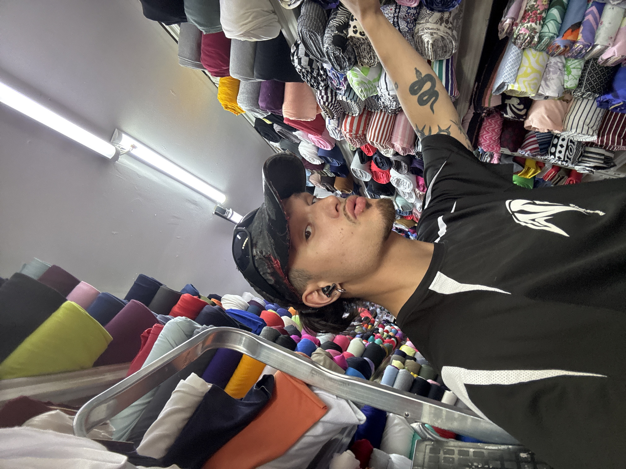 The image captures a young person with dark hair, a black baseball cap, and a black t-shirt, standing in a shop filled with rolls of fabric. The person is wearing earbuds and has a snake tattoo on their right arm. They are looking towards the camera with their lips pursed. The background is dominated by shelves stacked high with colorful rolls of fabric, ranging in color from white and orange to blues, purples, reds, yellows, and greens, with some patterned fabrics visible. A metal ladder is positioned to the left of the person. The lighting appears to be from overhead fluorescent tubes. The scene is likely a fabric store, potentially located in a market area within Sofia, Bulgaria, given the context. The person is in the process of browsing or selecting fabric, as indicated by their proximity to the rolls and their outstretched hand.
