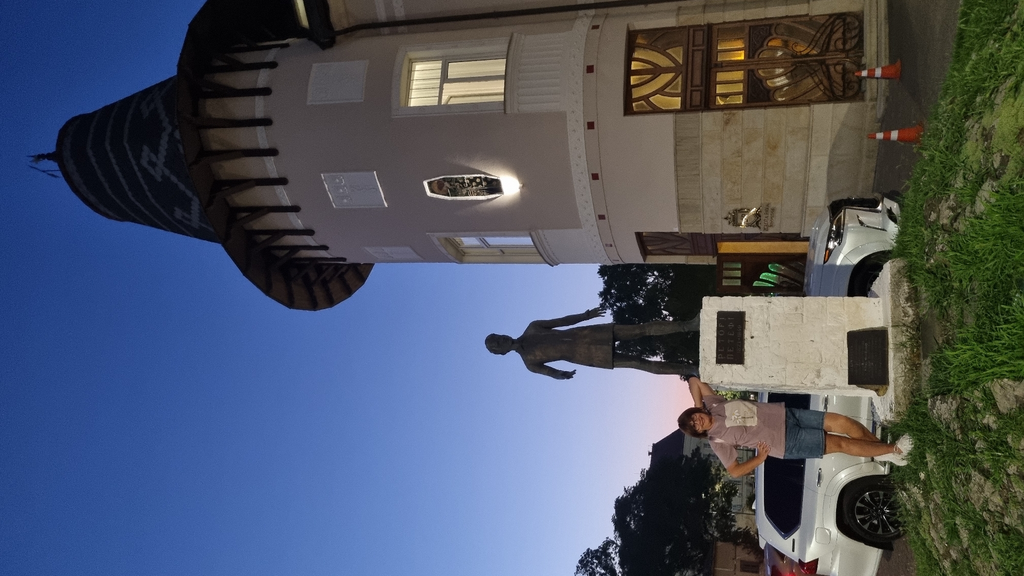 A woman stands next to a statue of a man in Berehove, Ukraine. The statue is on a stone pedestal, and there is a plaque on the pedestal. The woman is wearing a pink t-shirt and denim shorts, and she is leaning on the pedestal with her right hand. In the background, there is a building with a conical roof, and a white SUV. The sky is dark blue, indicating that it is either early morning or late evening. The statue is identified as Petőfi Sándor. The building in the background is the former Zrínyi Ilona High School, now the Zakarpattya Regional State Administration Building.