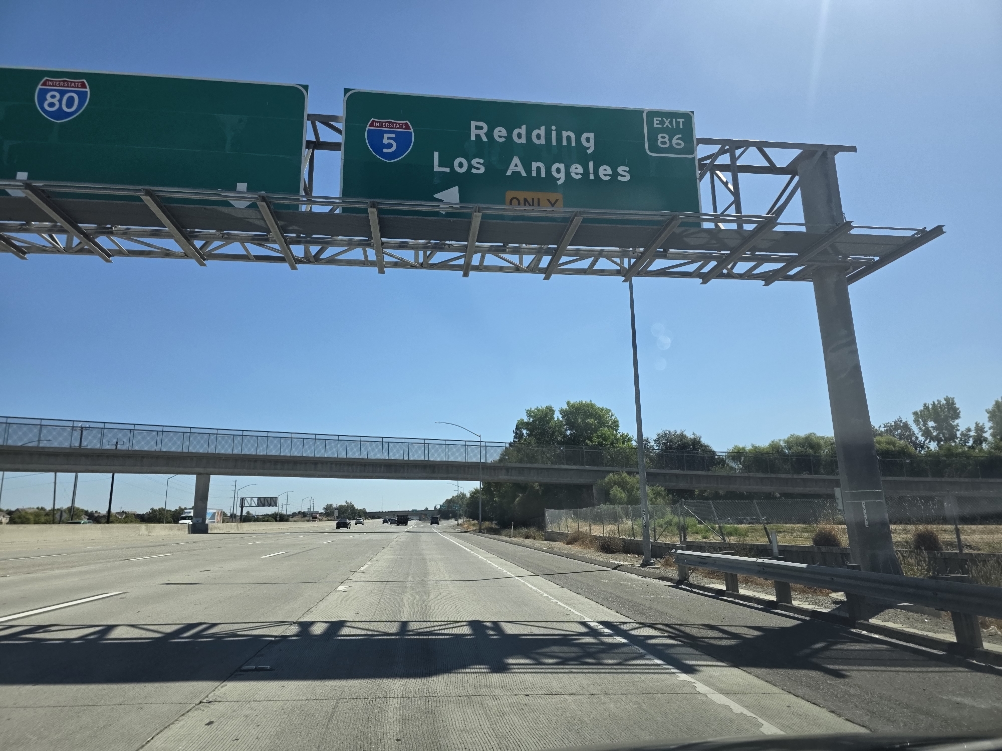 The image shows a view from a vehicle driving on a highway. Above the highway, a green overhead sign indicates "INTERSTATE 80" on the left and "INTERSTATE 5 Redding Los Angeles EXIT 86" on the right. A yellow sign below indicates "ONLY". The sky is blue and clear with some sun glare. In the distance, there are trees, other vehicles, and a pedestrian overpass. The highway appears to be in a suburban or rural area.