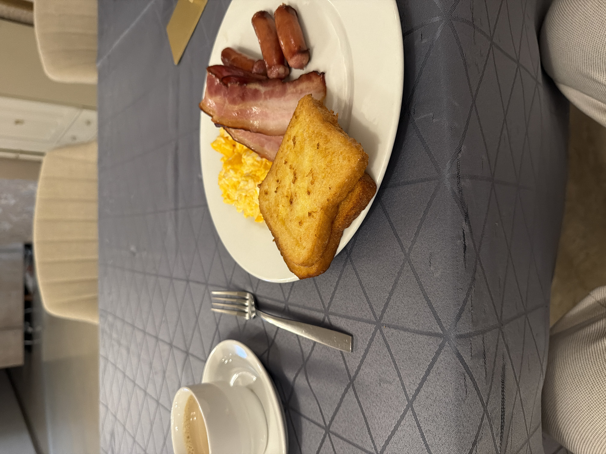 The image shows a breakfast meal on a table with a patterned, gray tablecloth. The meal consists of scrambled eggs, bacon, small sausages, and fried bread on a white plate. A fork is placed near the plate, and a cup of coffee with a saucer sits at the bottom of the frame. The scene appears to be indoors, possibly in a hotel or restaurant. The location of Pleven, Bulgaria is not explicitly evident from the image, but the meal could be served in a restaurant or hotel there.