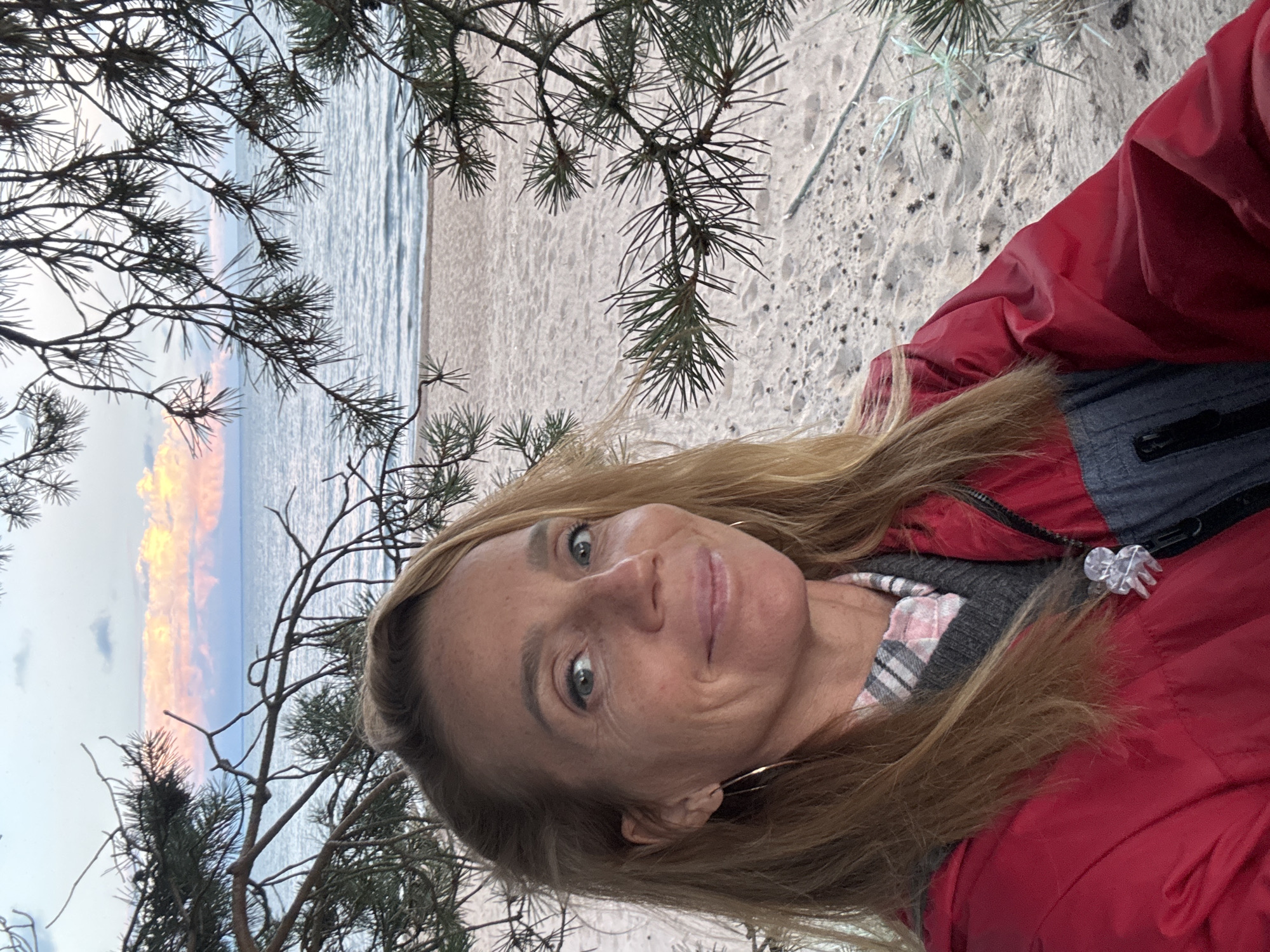 This is a close-up, selfie-style portrait of a Caucasian woman in her late 40s to early 50s, taken outdoors at a beach in Shepelevo, Russia. She has blonde hair, fair skin, and appears to be smiling slightly. She is wearing a red jacket over a grey sweater and a pink and white plaid shirt. A clear, abstract pin is attached to the left side of her jacket. The background shows a sandy beach, the sea, and a pink and orange sky at dusk, with pine branches framing the top of the image. The lighting suggests it is late evening.