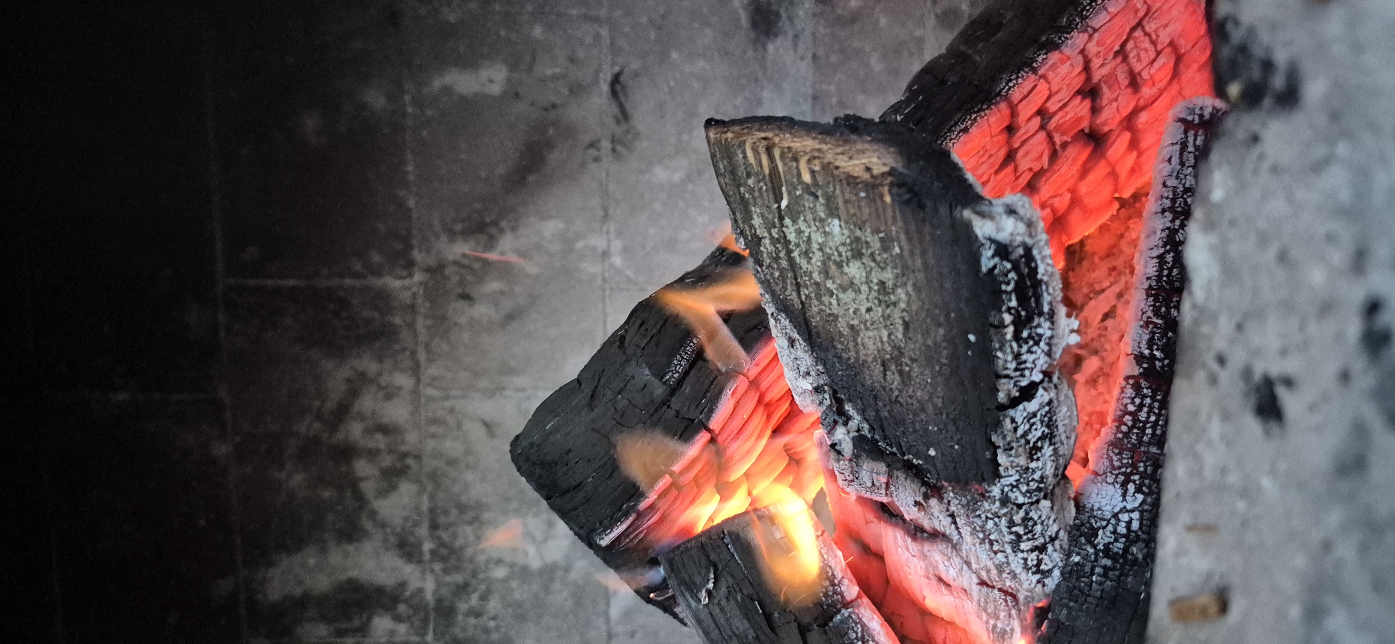 The image shows a close-up view of burning logs in a fireplace. The logs are charred black, with glowing red embers visible between them. Small flames flicker around the wood. The background consists of a grey, textured stone wall. The scene appears to be indoors, likely in a residential setting. The city of Ivancea, Moldova, is mentioned in the prompt, but there is no visual information in the image to confirm or deny this location.