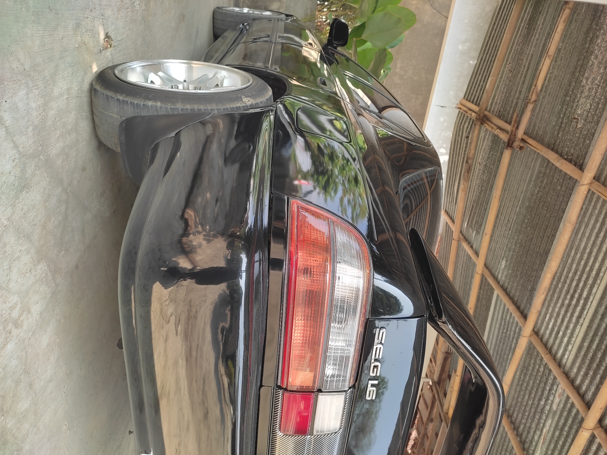 A black sedan, possibly a Toyota Corolla AE101, is parked on a concrete surface beneath a corrugated metal roof supported by bamboo poles. The car is viewed from a low angle, focusing on its rear and rear-left quarter. The vehicle has a spoiler on its trunk and a silver emblem that reads "SEG 1.6". The taillights are a combination of clear, amber, and red. The rear tire is silver with five spokes. A large green plant is visible to the left of the car. The background shows a wall and the roof structure. The scene appears to be an outdoor parking area in Gandu, Indonesia, likely a residential or semi-rural setting.