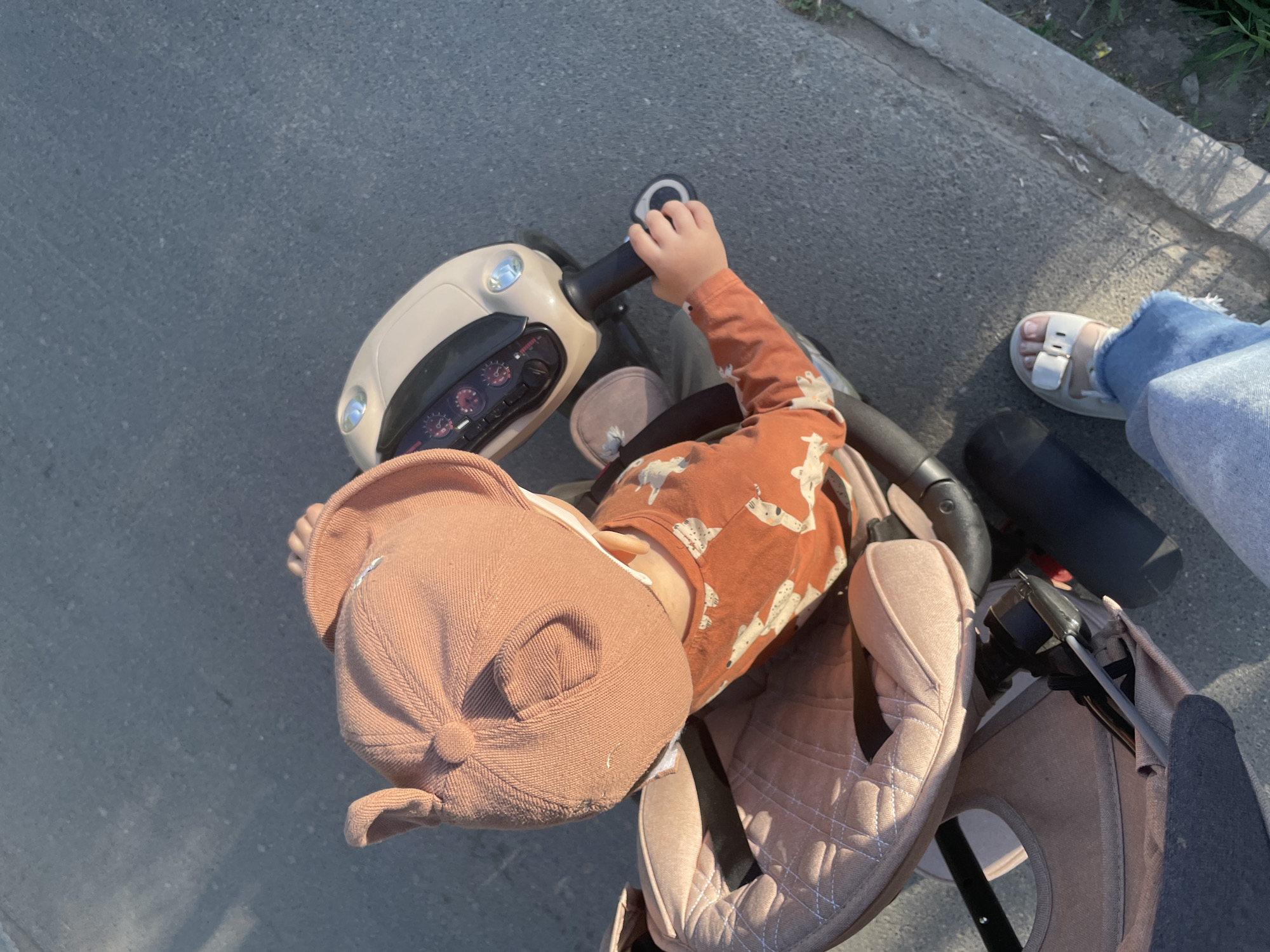 A high-angle shot depicts a child riding a light-colored tricycle. The child wears a peach-colored hat and a patterned, long-sleeved, reddish-brown shirt. Their right hand holds the tricycle's black handlebars. Next to the tricycle, a leg clad in blue jeans and a white sandal is visible. The setting appears to be outdoors on a paved surface.
The image does not contain the necessary information to place it in Izhevsk, Russia.