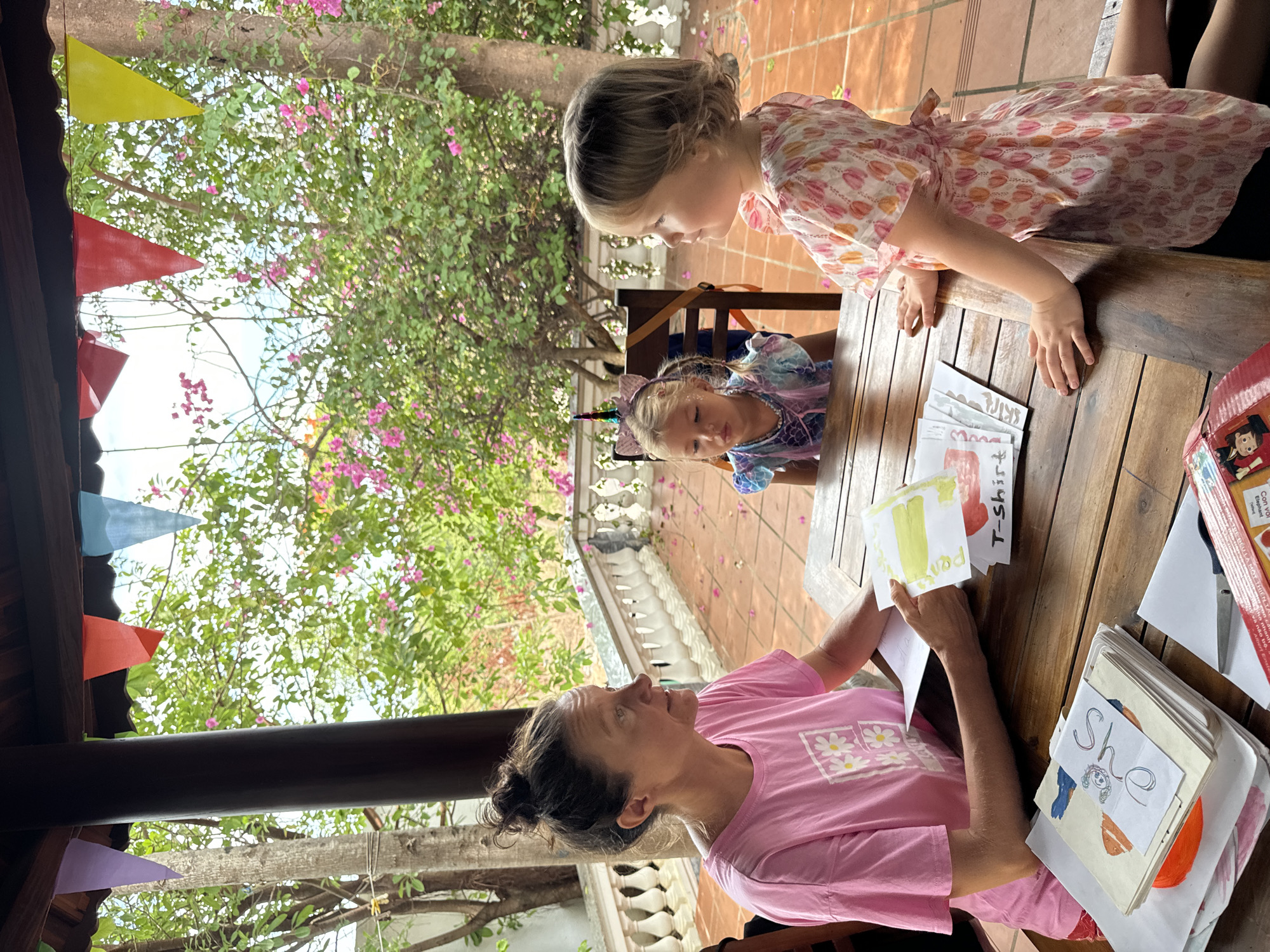 An adult female in a pink t-shirt sits at a wooden table with two young girls under an outdoor canopy decorated with colorful triangular flags. One girl, wearing a floral dress, leans over the table, while the other, sporting a unicorn headband, sits on the opposite side. The table is covered with educational materials including illustrated flashcards and handwritten notes. The scene is outdoors, likely in Mũi Né, Vietnam, evidenced by the location description in the corner of the picture. The background features a lush green tree with pink blossoms and a tiled patio. The woman appears to be teaching or interacting with the children using the educational materials.