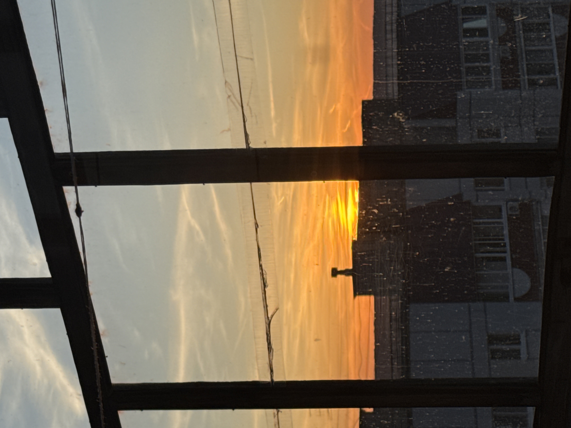 The image shows a sunset view through a window frame, possibly in Chaikovsky, Russia. The sky transitions from light blue to orange and yellow, reflecting off the glass. A building is visible in silhouette on the right, showing window patterns and architectural details. The window frame itself is made of dark material, and there are several support structures that divide the scene. The photograph is taken at an angle.