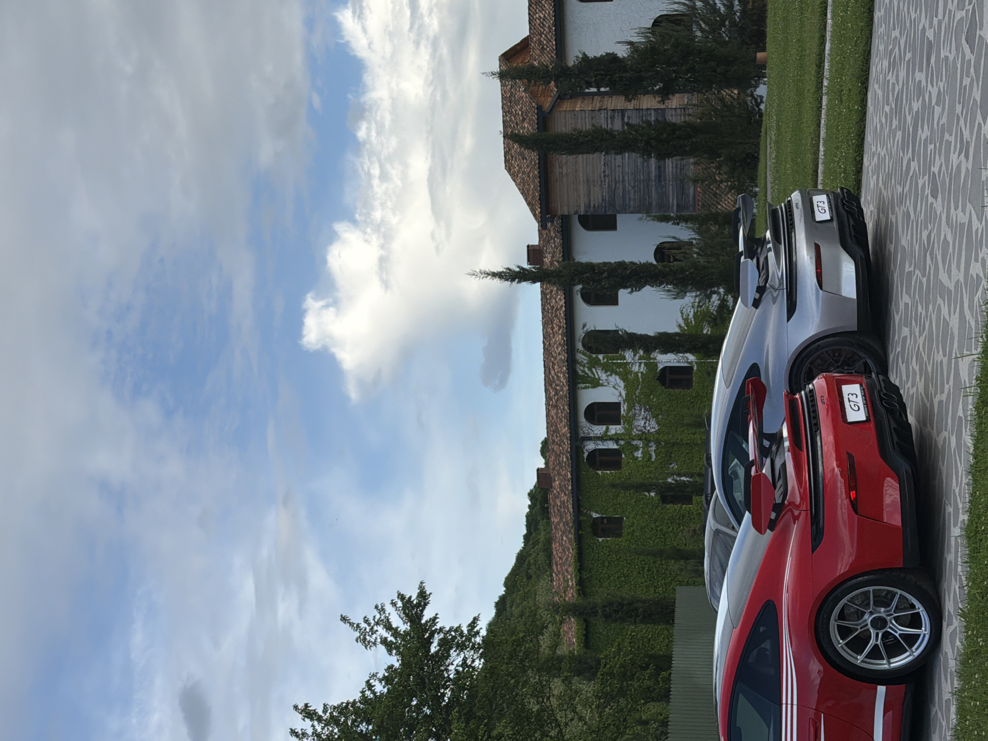 The image shows two parked Porsche GT3 cars, one red and one silver, in what appears to be the city of Napareuli, Georgia. The cars are parked on a stone-paved area next to a grassy lawn. Behind the cars is a building covered in ivy, with traditional architectural details. The sky is partly cloudy. The image seems to be taken on a sunny day.