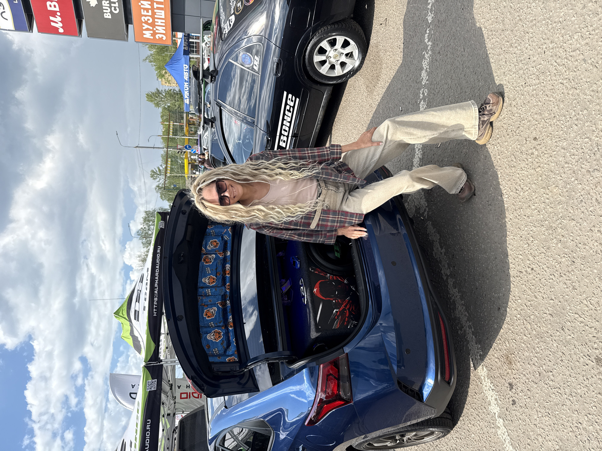 A medium shot captures a woman with long, blonde, wavy hair perched on the trunk of a blue car. She sports sunglasses, a flannel shirt, and light-colored pants. The car trunk is open, revealing sound equipment adorned with tiger-print fabric.
Beside the blue car is another car, black, parked on the asphalt ground of what appears to be a car show or outdoor event. In the background, various signs are visible, including ones advertising "M.B," "BUR," "CLU," "МУЗЕЙ ЭЙНШТЕ," and "ДИКОН АВТО." A white tent with the logo "AUDIO.RU" can also be seen. The location is in the city of Unknown, Unknown, but no specific details indicate a particular place within the city. The sky is overcast, suggesting the photo was taken on a partly cloudy day.