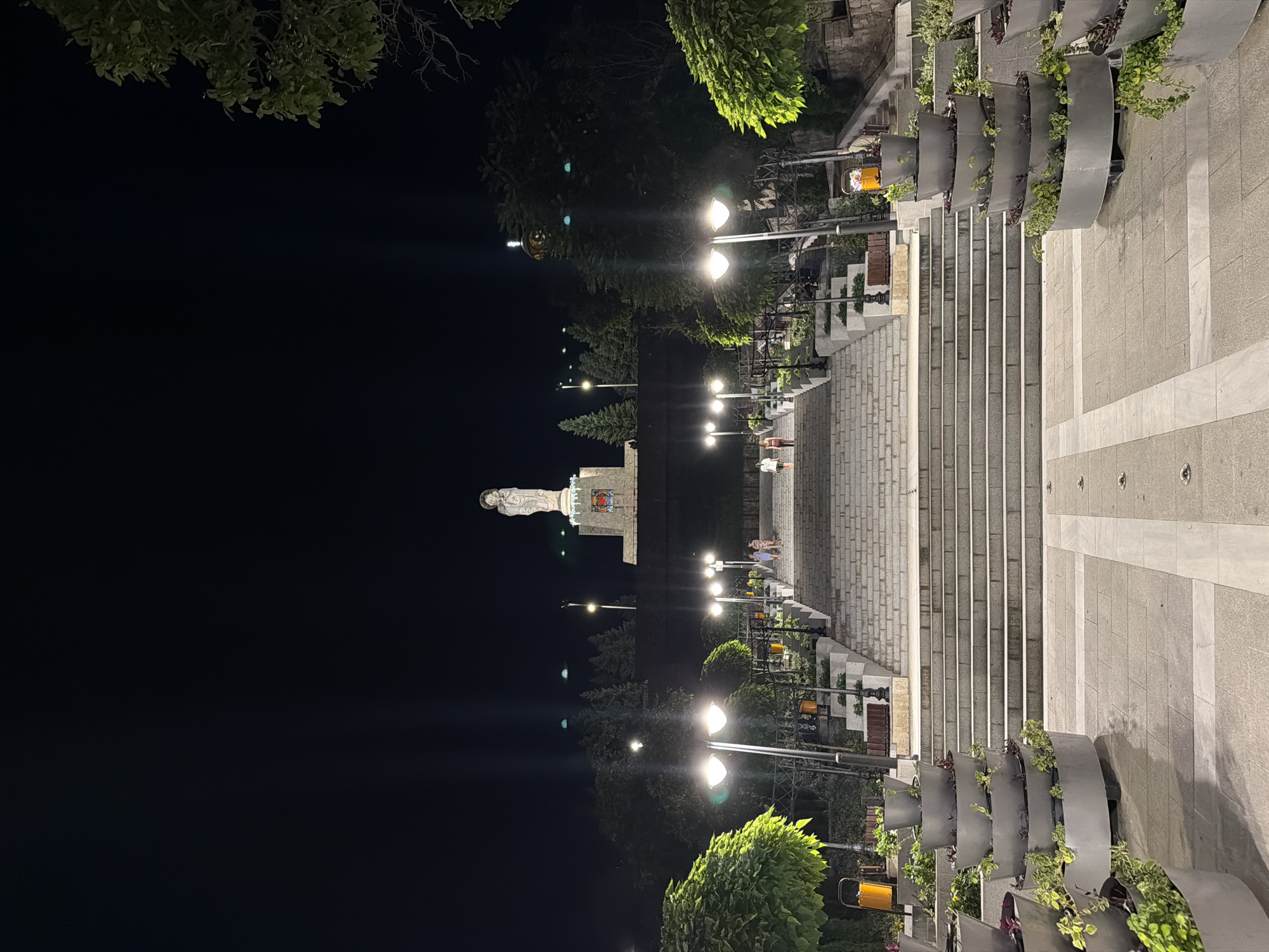 The image captures a nighttime view of the Monument of the Holy Mother of God in Haskovo, Bulgaria. A grand stone staircase ascends towards the monument, which is illuminated and stands atop a pedestal. Several streetlights line the staircase, casting a warm glow on the surroundings. Greenery, including trees and potted plants, is visible on either side of the stairs. A few people are seen walking up the stairs, and a statue of the Virgin Mary holding a child is prominently displayed at the top. The scene is set against a dark night sky.
