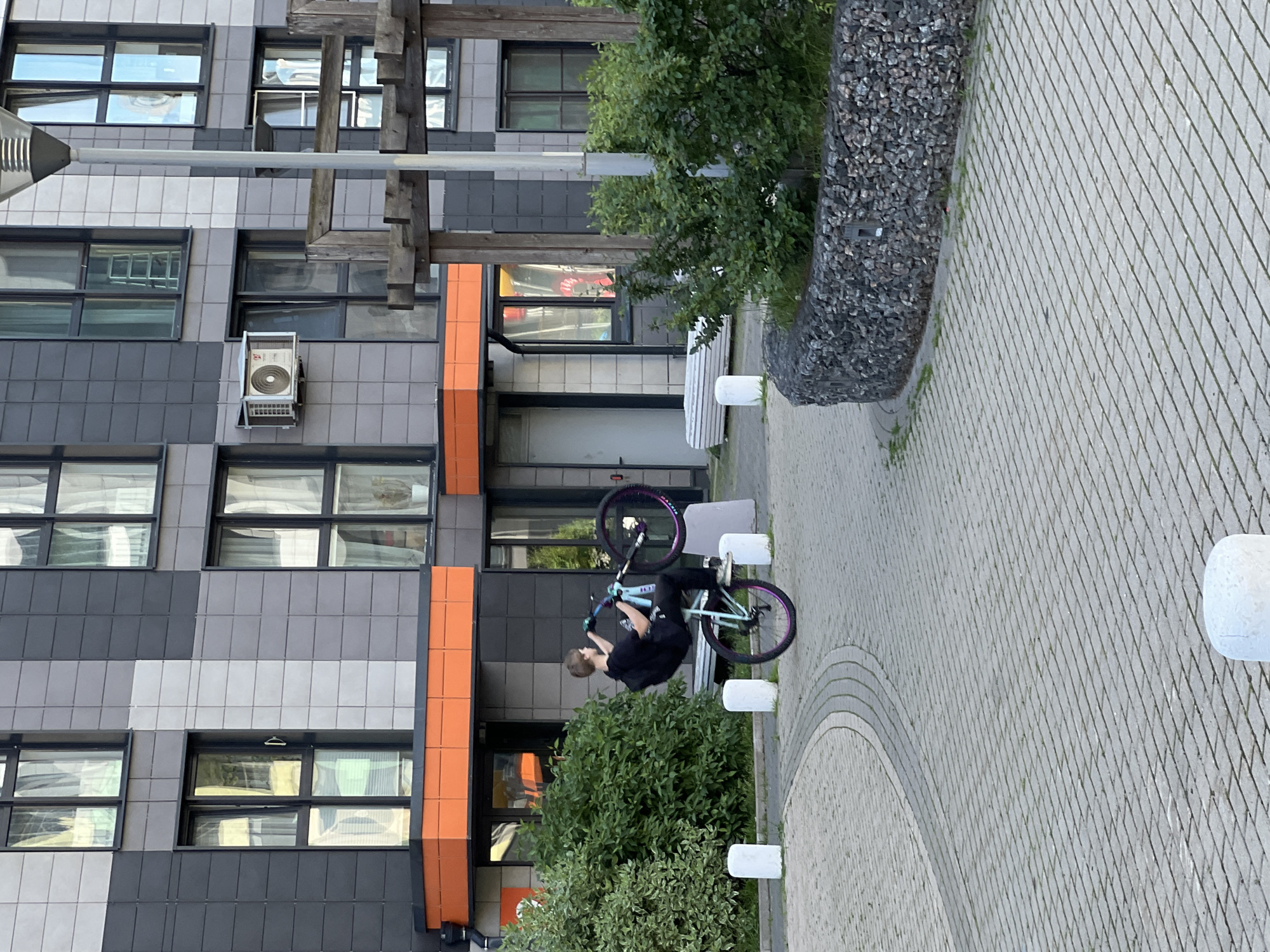 The image depicts a young person performing a stunt on a BMX bike in an urban courtyard. The person, dressed in black, is mid-air, with the bike positioned vertically. The scene is set in a modern residential complex in Mytishchi, Russia, characterized by its modular facade featuring a mix of grey, white, and orange tiles. Large windows are visible on the building. The ground is paved with interlocking concrete blocks forming a patterned walkway. A gabion wall filled with dark stones and lush green bushes frame the courtyard. White cylindrical bollards are placed along the walkway. The lighting suggests it's daytime, likely afternoon, with soft shadows indicating overcast or diffused sunlight. The overall impression is one of dynamic action within a contemporary urban environment. No visible text is present in the image.