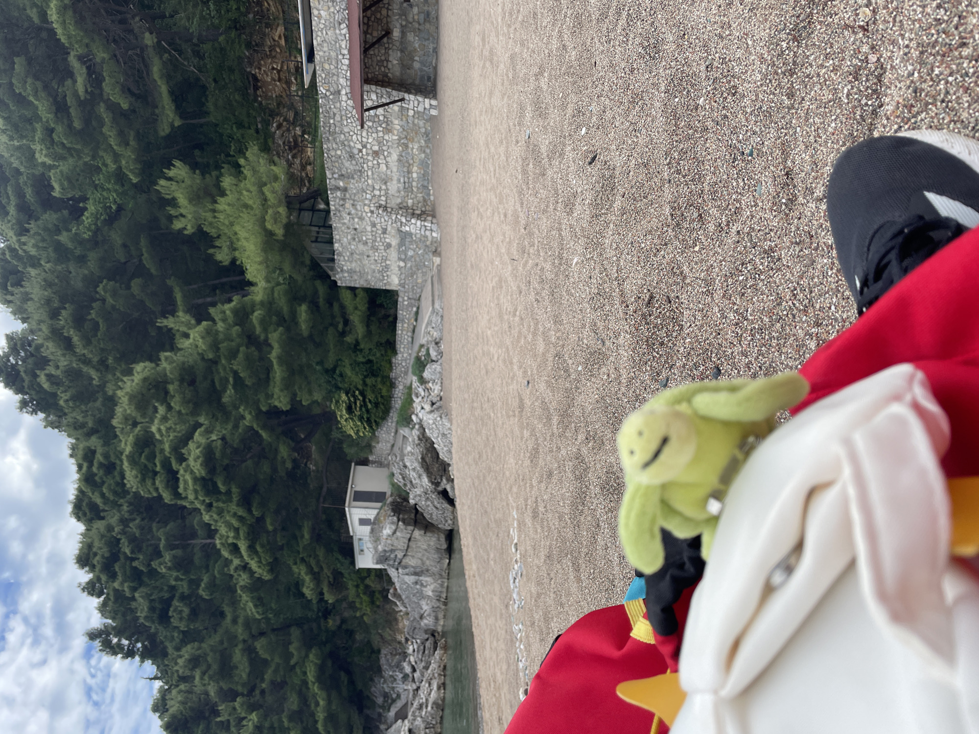 The photograph, taken at an angle, depicts a beach scene in Bečići, Montenegro. The foreground shows a person's lap wearing red clothing, with a black and white sneaker visible. Two plush toys, a green frog and a white figure, are partially in view. The middle ground displays a sandy beach extending towards a structure made of stone, likely a building or fortification. Lush green trees and vegetation cover the left side of the image, descending towards the water's edge, where a small white building is perched on the rocks. The sky is partly cloudy.