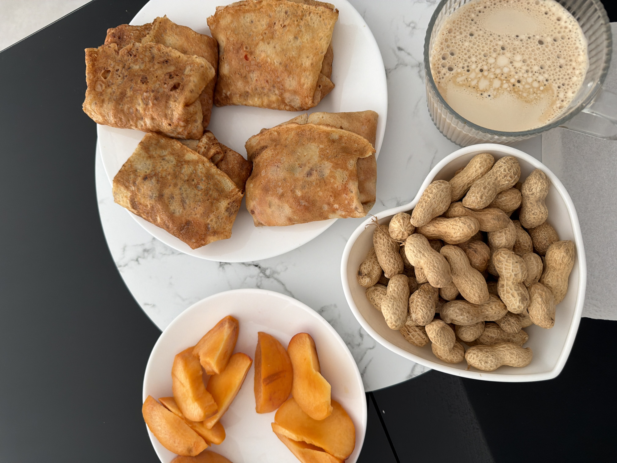 An overhead shot shows a breakfast spread on a black and white marbled tabletop. In the foreground, a white heart-shaped bowl overflows with peanuts in their shells. To the right, a clear ribbed glass is filled with a frothy beverage, possibly coffee with milk. Behind the peanuts and glass, a white plate holds four golden-brown crepes, folded and stacked. To the left of the crepes, a white plate contains several bright orange slices of fruit, likely persimmon or peach, cut into wedges. The scene is lit from above, casting subtle shadows. The image is an indoor shot, likely taken in a kitchen or dining area, with no visible exterior elements or indications of the specific city of Finestrat, Spain.