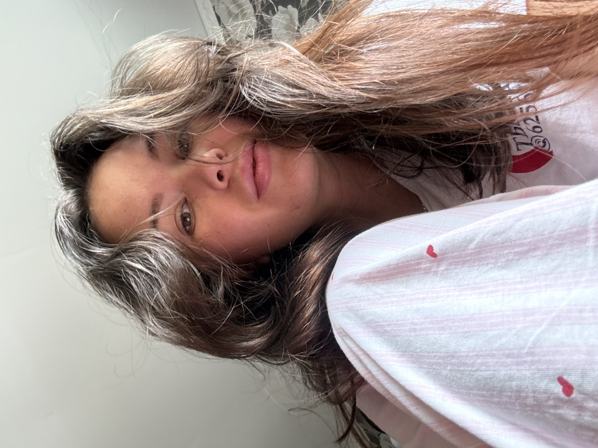A close-up, low-angle, eye-level shot shows a young adult woman with long, dark brown hair. She is wearing pink and white striped pajamas with small red hearts. Her hair is messy and partially obscures her face, with strands falling across her eyes. She has fair skin and is looking slightly away from the camera. The background is a blurred wall with a floral pattern. The overall lighting suggests an indoor setting, likely a bedroom. The image was taken in Bely Yar, Russia.