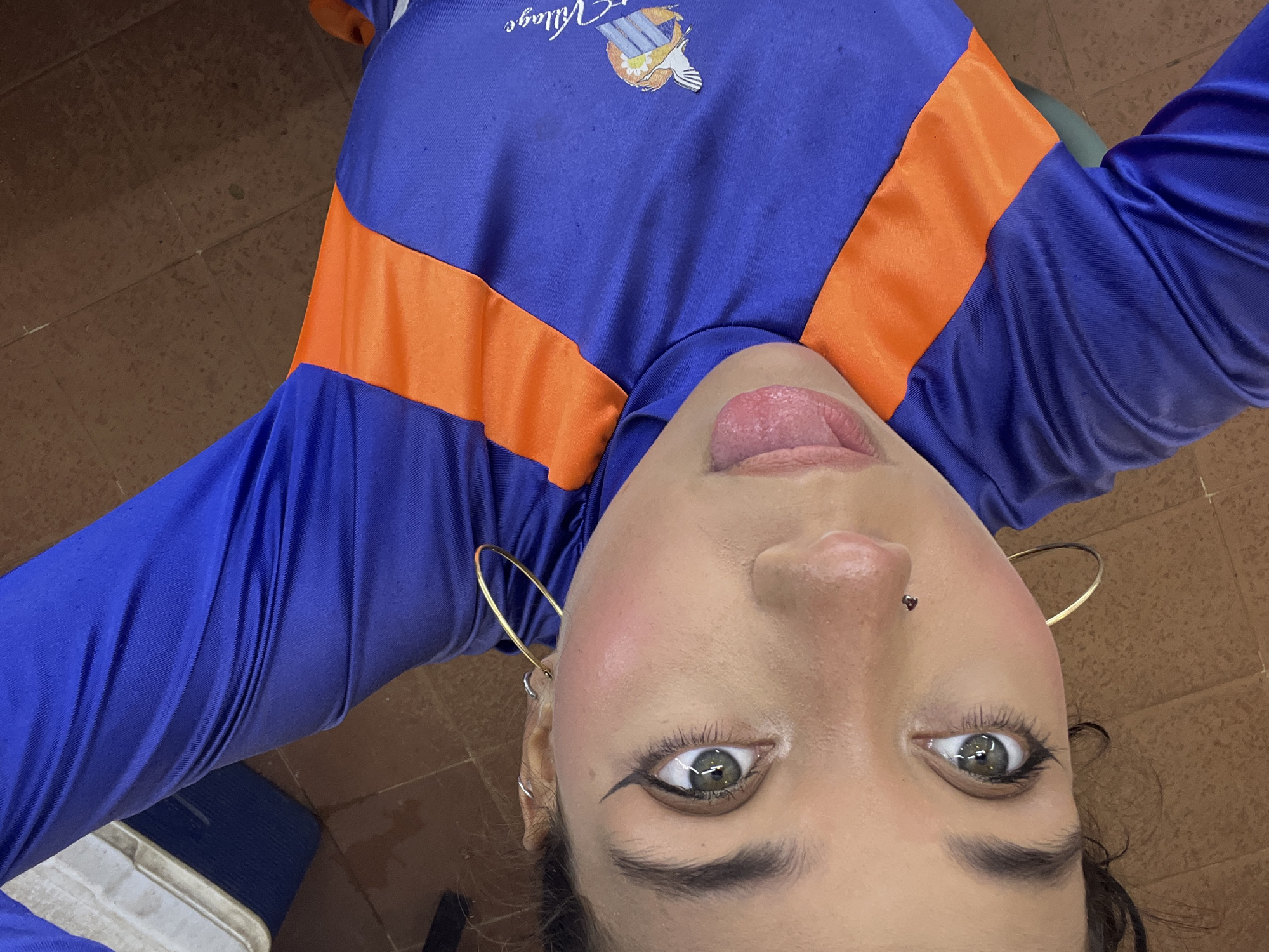 The image shows a close-up, overhead view of a young woman lying down, likely on a tiled floor, with her head tilted and facing the camera. She is wearing a blue and orange long-sleeved shirt. Her eyes are open, her tongue is slightly extended, and she has dramatic winged eyeliner. She is wearing large hoop earrings and a small stud piercing on her nose. The overall impression is of a playful or candid moment. The woman's clothing features a logo on the right side of the shirt that appears to be for "Village." There is a cooler visible in the upper left corner of the image. The floor is tiled with reddish-brown squares. There is no discernible location in the city of Porlamar, Venezuela, that can be identified from this image.