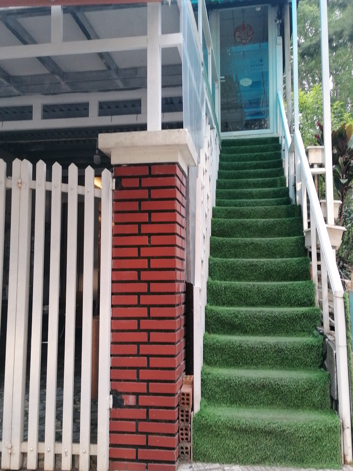 A view of a staircase covered in artificial grass leads up to a glass door of a building. The staircase is flanked by white railings and a brick pillar on the left. A white picket fence is positioned next to the brick pillar. The scene appears to be outdoors, possibly in a residential or commercial area in Da Lat, Vietnam.