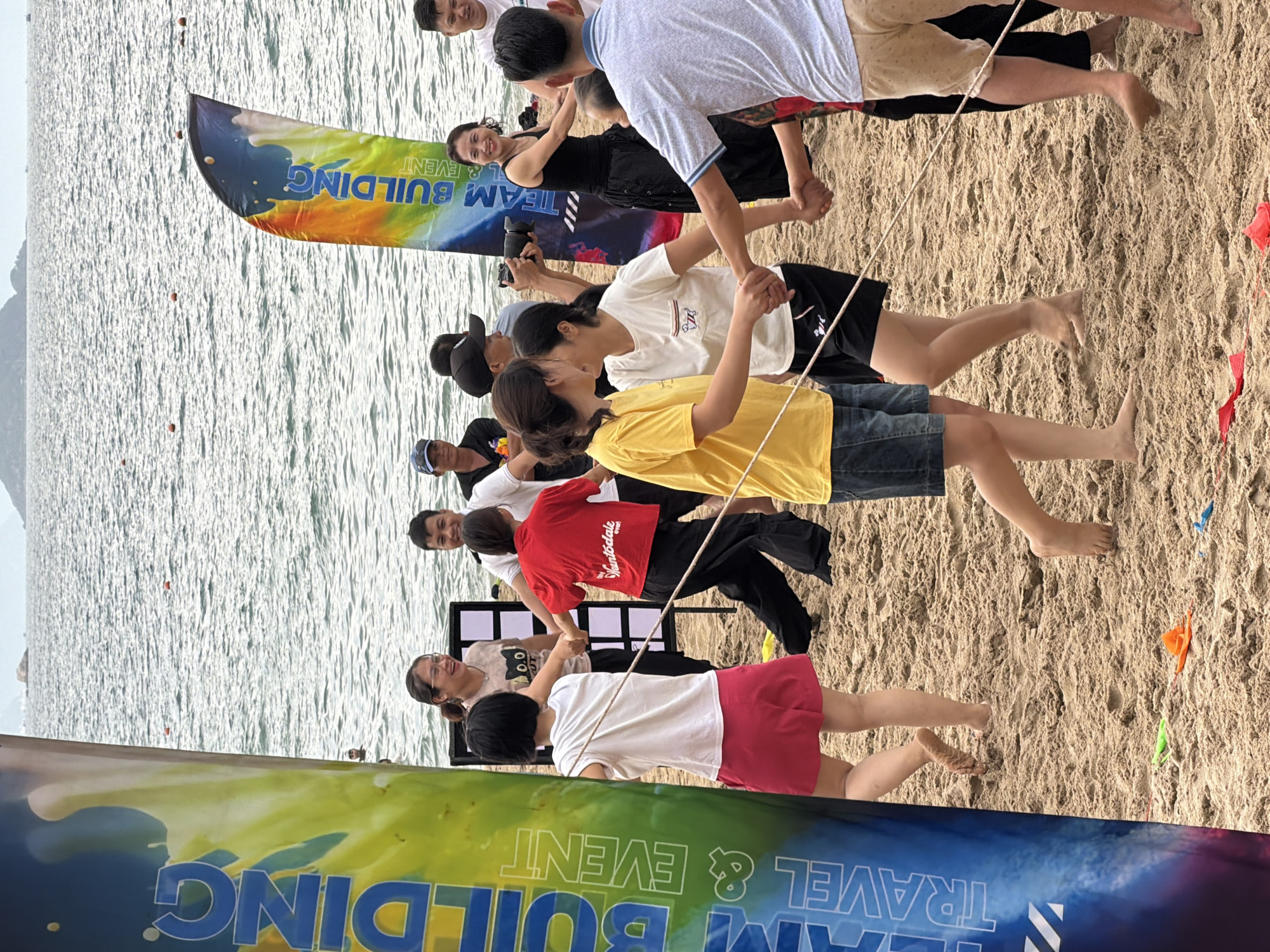 The image depicts a group of people participating in a team-building activity on a sandy beach in Nha Trang, Vietnam. The background features the sea with a distant island or coastline. A large, colorful banner on the left side of the frame has text that partially reads "TEAM BUILDING TRAVEL & EVENT." Another similar banner is visible on the right. In the foreground, several individuals are holding hands in a circle, suggesting they are engaged in a game or activity. Some are smiling and looking at each other, indicating interaction and engagement. The people are dressed casually in t-shirts, shorts, and skirts. The sand appears damp in some areas, and there are some colorful plastic objects scattered on the ground, possibly part of the activity. The overall atmosphere appears to be one of fun and camaraderie.