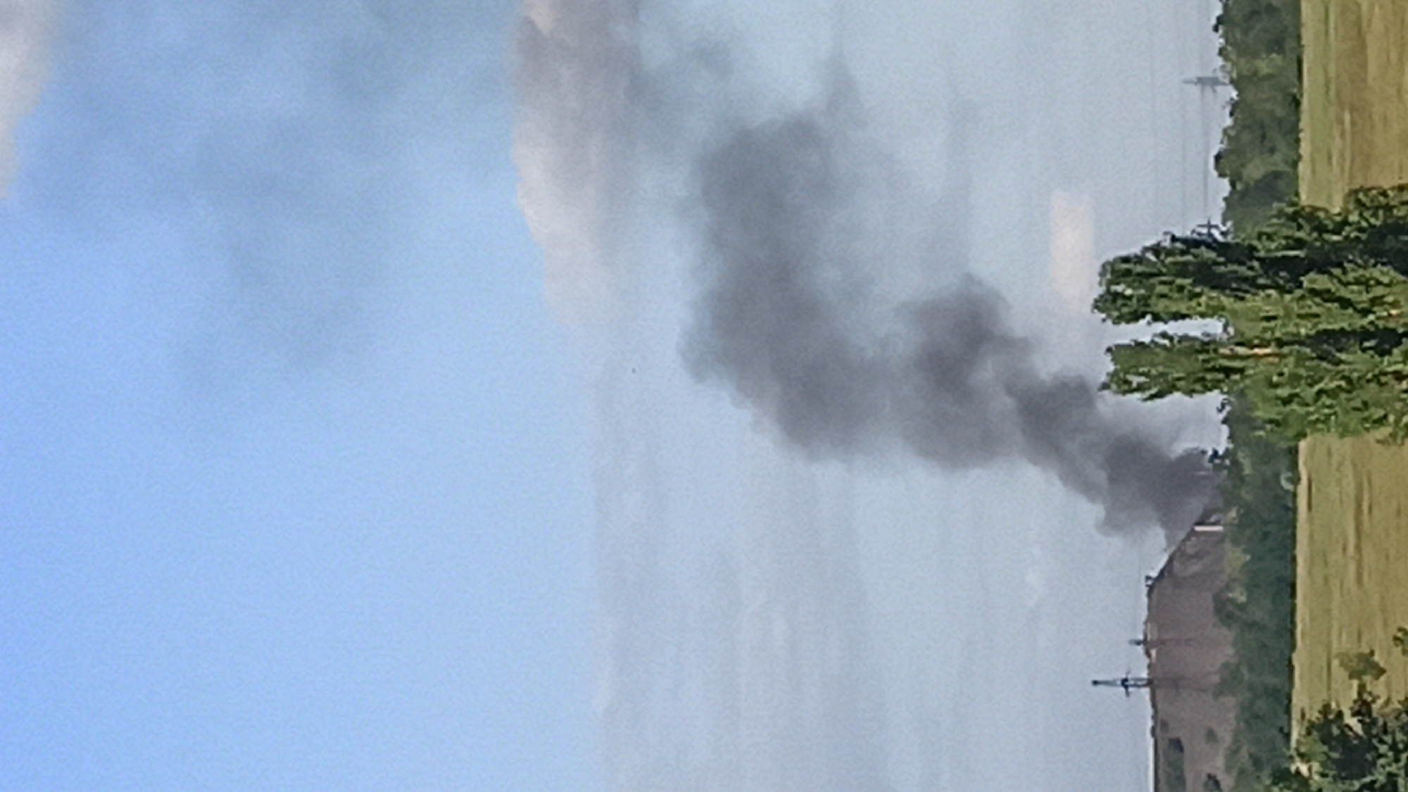 In the city of Unknown, Unknown, a plume of dark smoke billows into a pale blue sky with scattered clouds. In the mid-ground, a large slag heap or industrial mound dominates the left side of the frame, with a tall industrial structure on its peak. The smoke appears to originate from the base of this mound, obscuring some of the industrial activity there. In the foreground, lush green trees and fields create a natural barrier, with two tall, slender trees with dense foliage prominently placed in the right mid-ground. The overall impression is an industrial landscape juxtaposed with natural elements under a hazy sky.