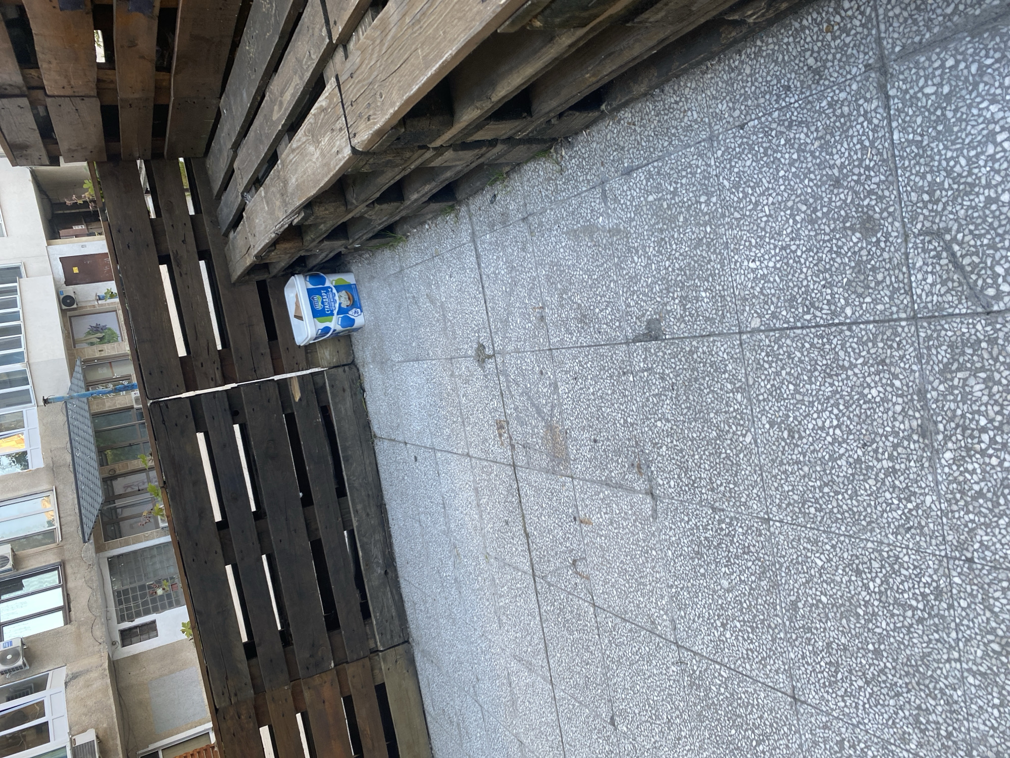 A medium shot depicts a portion of a building facade on the left, a stack of wooden pallets on the left and top, and a tiled ground surface in Sofia, Bulgaria. In the center, a cylindrical container with blue and white labeling is present. The building has multiple windows and some air conditioning units. The pallets are weathered, with gaps between the wooden slats. The tiled ground has a fine, speckled pattern.