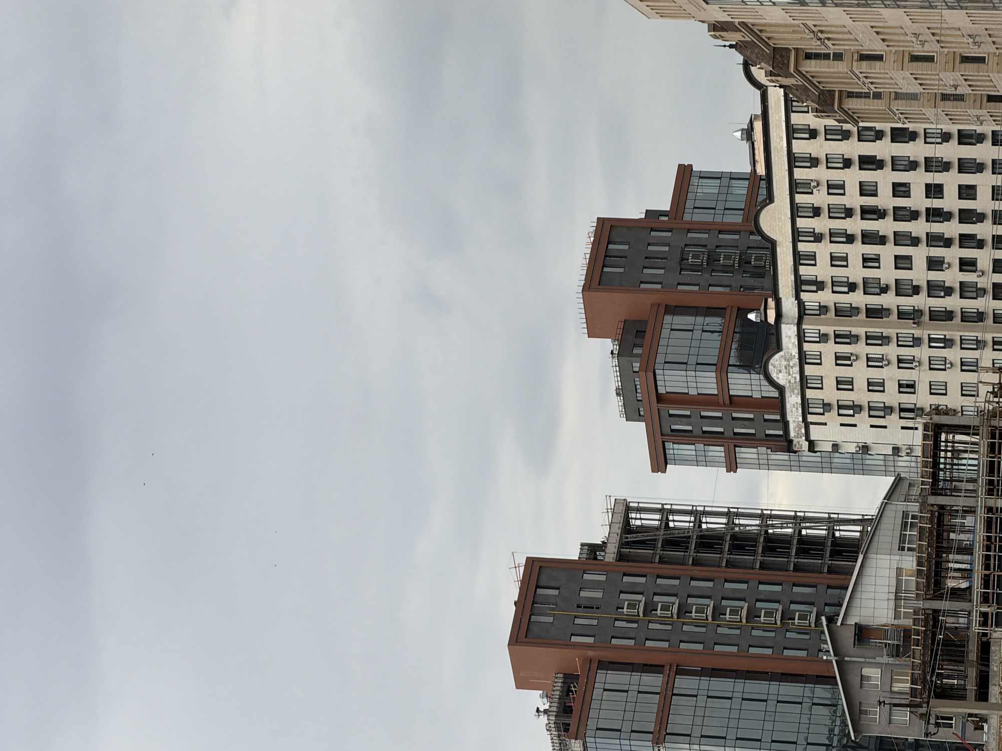 A low-angle, full shot captures a cluster of modern buildings against a heavily overcast sky. The buildings exhibit a mix of architectural styles and stages of construction. In the foreground, a multi-story structure with exposed rebar and concrete framing indicates an ongoing construction project. Adjacent to this, a gray, low-rise building with multiple windows and a prominent central door appears completed.
Dominating the mid-ground are two taller, contemporary buildings. The one on the left features dark gray facades punctuated by numerous windows and accented with reddish-brown trim. This building also shows signs of ongoing work, with visible scaffolding and exposed structural elements on its upper floors. To its right, a similar building with a dark gray core and reddish-brown framing rises, featuring large glass sections.
Further to the right, a lighter-colored building with a repeating pattern of windows and a mansard roof stands out. Its facade is detailed with ornate architectural elements. The rightmost building, partially visible, is also light-colored with a similar repeating window design. The overall scene suggests a developing urban landscape under a muted, cloudy sky, with varying levels of completion evident across the structures. There are no people or animals visible in the frame.