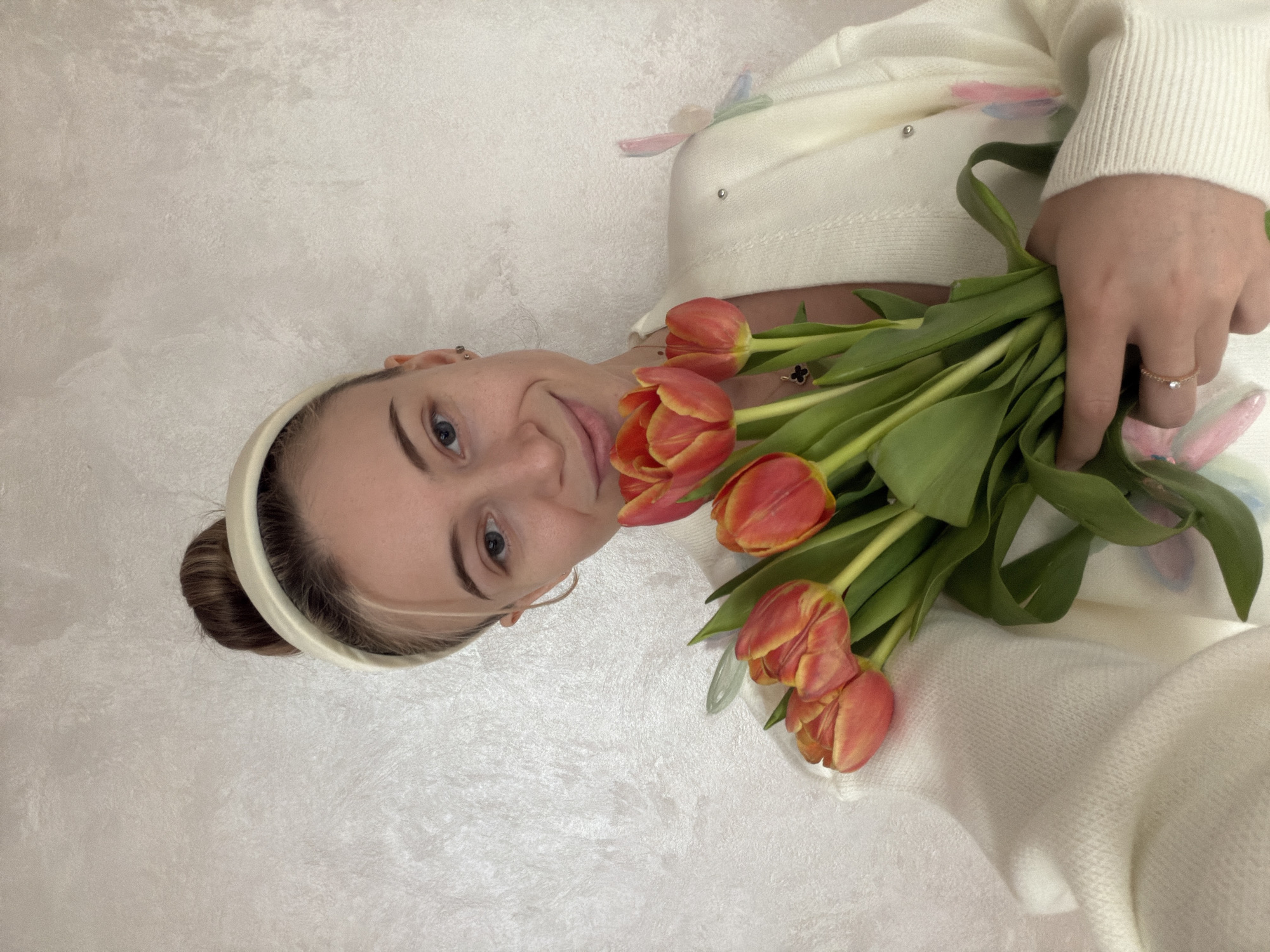 A woman is shown from the chest up, holding a bouquet of orange and yellow tulips. She has fair skin, blue eyes, and her hair is pulled back into a bun, secured by a cream-colored headband. She is wearing a cream-colored cardigan with small pearl-like embellishments. She is also wearing a delicate gold ring on her ring finger. The background appears to be a textured off-white wall. The location is unknown, and not confirmed as Plovdiv, Bulgaria.