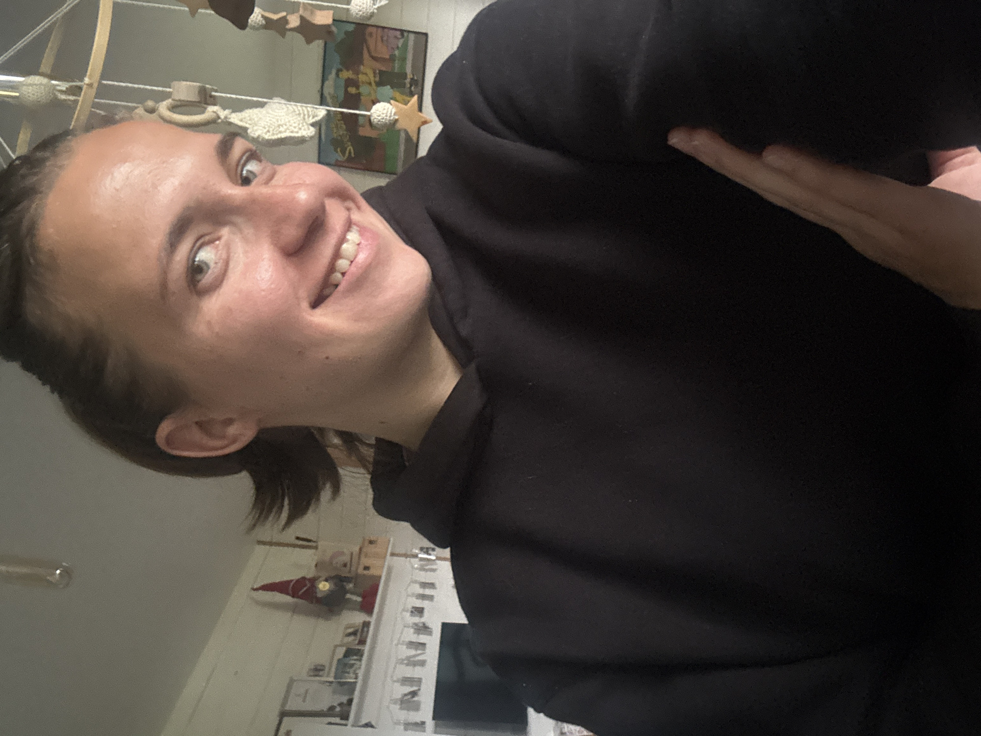 A person wearing a black hoodie is smiling at the camera. The location appears to be indoors, with a decorated shelf and a baby mobile visible in the background. It is not possible to confirm the specific location of Раздолье, Russia.