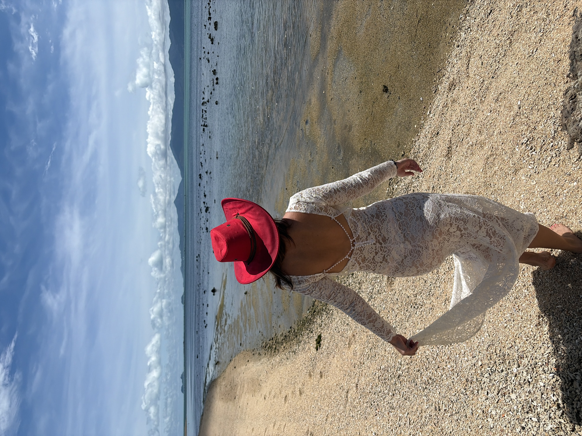 A woman wearing a white lace dress and a red hat stands on a sandy beach in Baan Bo Phut, Thailand. She is facing away from the camera, looking towards the ocean. The beach is made of small pebbles and sand, with the water's edge visible in the foreground. The ocean is calm with a few scattered rocks and islands in the distance. The sky is partly cloudy with a bright blue hue. The woman is holding the hem of her dress, which has long, sheer sleeves. Her hair is dark and her back is adorned with a string of pearls.
