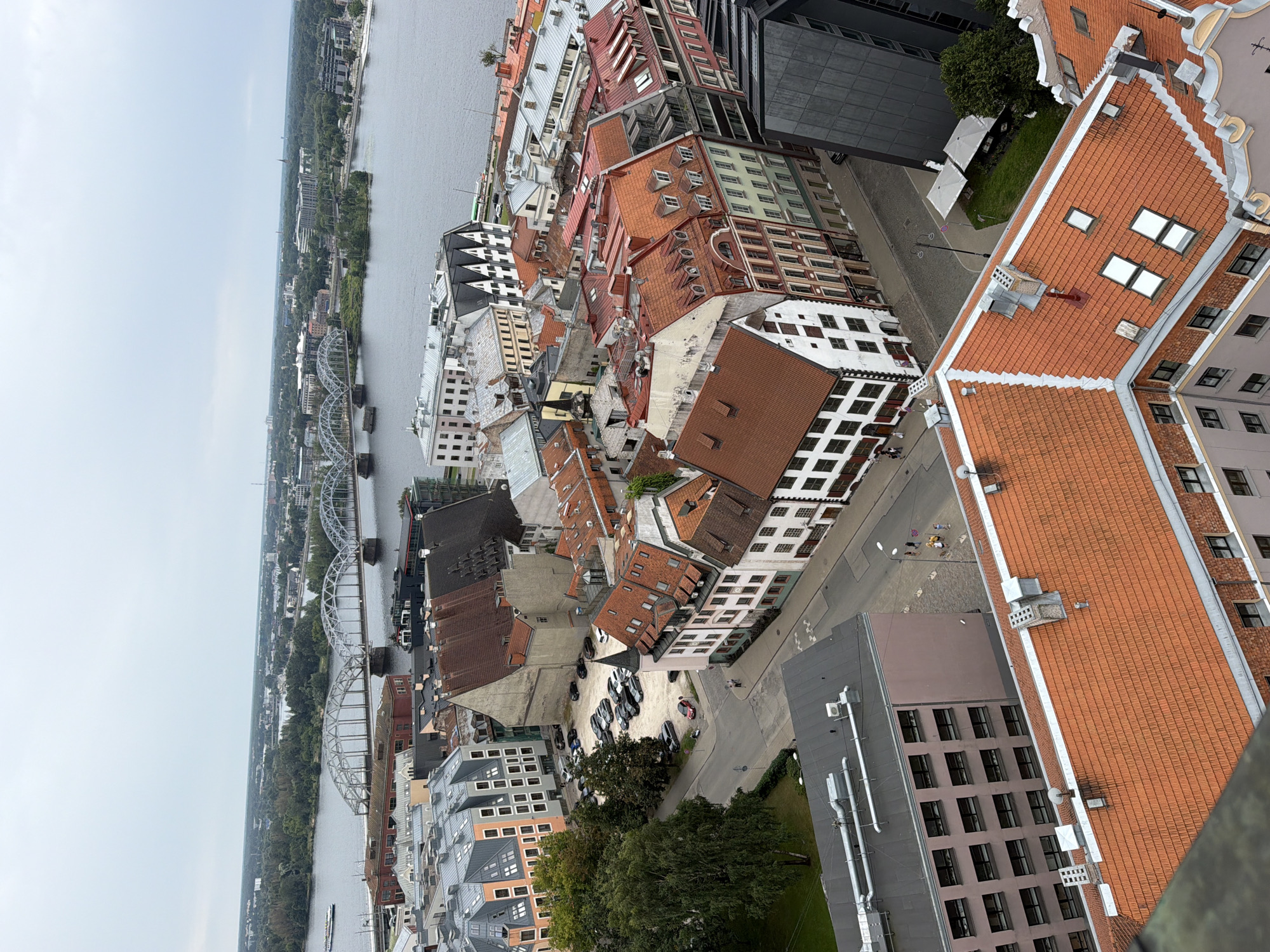 This is an aerial view of the city of Riga, Latvia, capturing a section of the Daugava River and a prominent railway bridge. In the foreground, a cluster of buildings with red-tiled roofs dominates the scene, showcasing a mix of architectural styles. To the left, a parking lot filled with cars is visible amongst lush green trees. The river flows calmly, with a boat visible in the distance. The background reveals a cityscape with more buildings and dense greenery, extending towards the horizon under a cloudy sky. The overall impression is that of a historic European city, with a blend of old and new architecture, and the presence of a major waterway shaping its landscape.