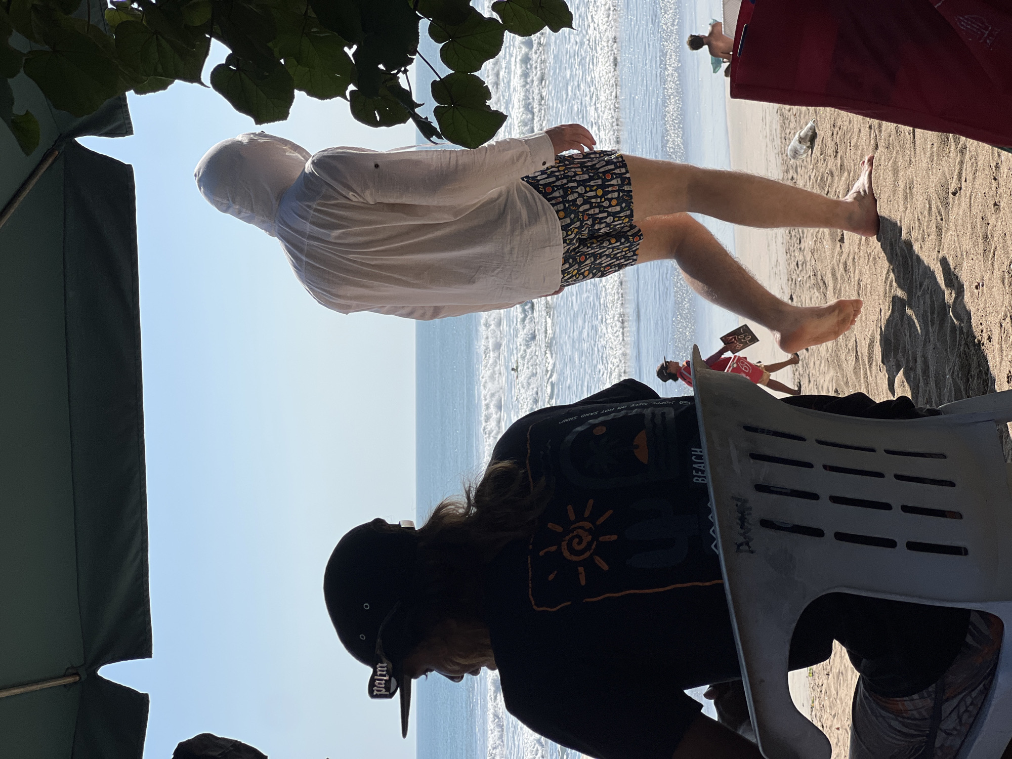 A sunny day at Kuta Beach, Indonesia, showcases two individuals. In the foreground, a person wearing a black t-shirt with an orange sun graphic and a black baseball cap with "palm" written on it sits in a white plastic chair, facing away from the camera. To their right, a person in a white hooded shirt and patterned shorts walks away from the camera towards the ocean. The beach is sandy, with gentle waves crashing on the shore. Other beachgoers are visible in the distance. The scene is partially shaded by a large green umbrella and foliage.