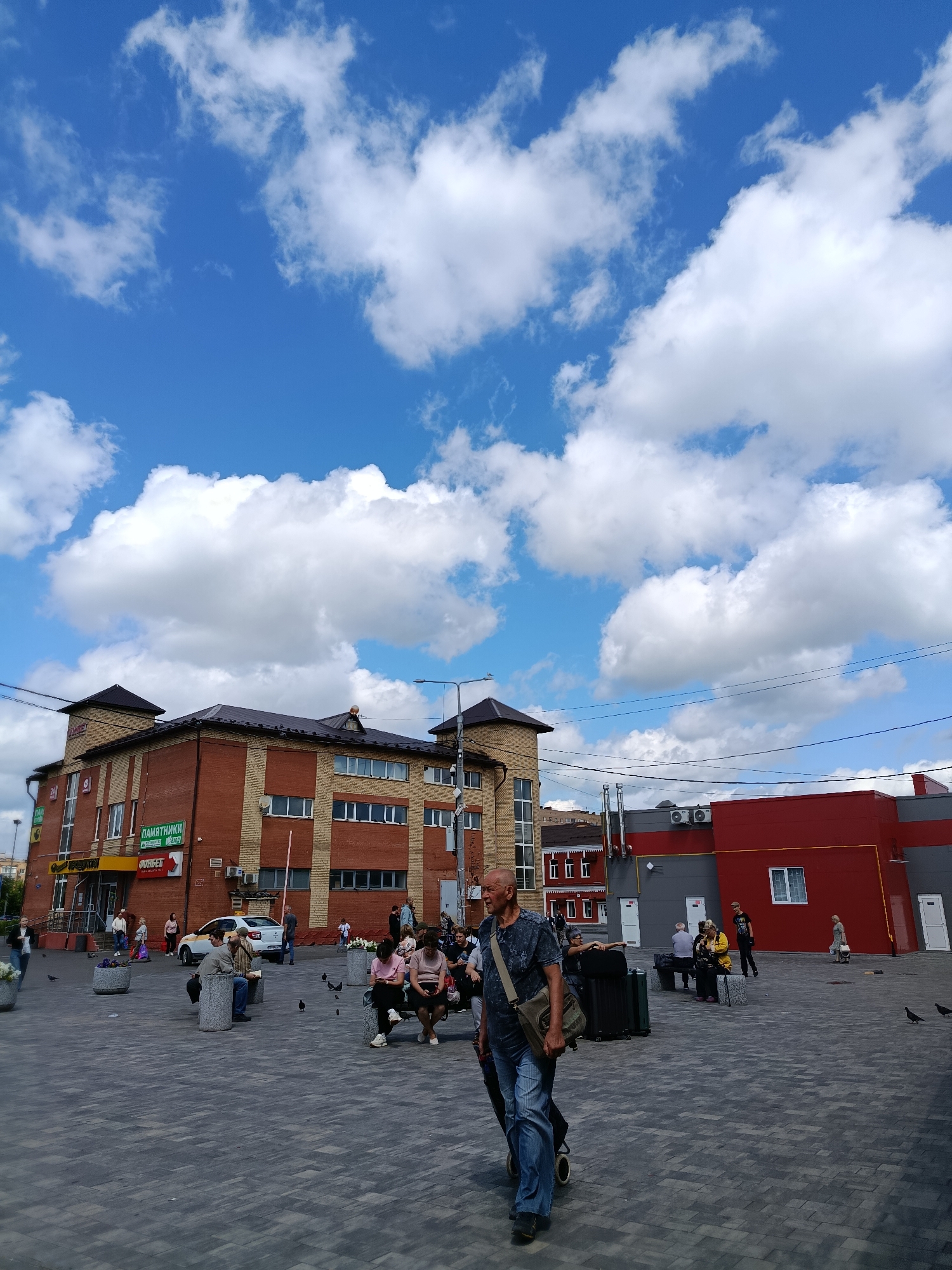 In a city square in Orekhovo-Zuyevo, Russia, under a bright blue sky with scattered white clouds, a man in a dark t-shirt and blue jeans walks towards the camera, carrying a messenger bag. To his left is a red building with gray accents, and beyond that, a multi-story brick building with dark roofs and many windows. Several people are seated on benches or standing in the square. Some are looking at their phones, while others appear to be conversing. A white car with a yellow stripe is parked near the brick building, and several pigeons are scattered across the paved ground. The overall scene depicts a sunny day in an urban public space with pedestrian activity.