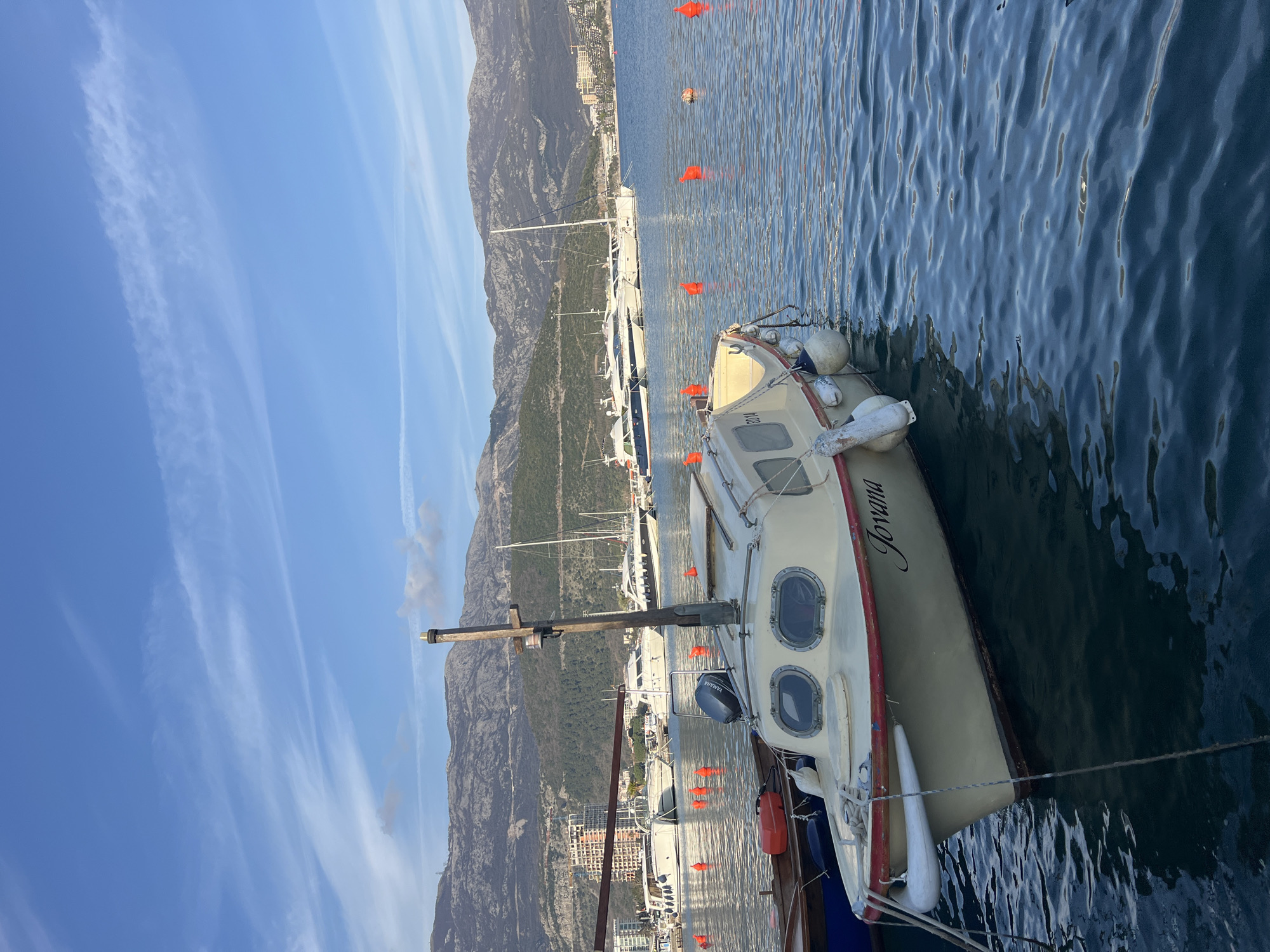 The image depicts a marina scene in Budva, Montenegro, under a clear blue sky with wispy clouds. In the foreground, a light-colored motorboat named "Jovana" is moored, displaying a red trim along its hull, two oval portholes on its cabin, and multiple white cylindrical fenders. A blue Yamaha outboard motor is visible at its stern, and a wooden mast-like pole with a light fixture stands on the cabin. Ropes secure the boat. To its immediate left, a portion of another dark-hulled boat is visible. The water is calm with ripples reflecting light.
In the midground, numerous orange spherical buoys are distributed across the water, marking various points. Several larger white yachts and sailboats are moored further back in the harbor. The background features prominent, rugged, forested mountains, characteristic of the Montenegrin coastline. At the base of these mountains, along the coastline, several urban structures are visible, including multi-story buildings and what appears to be ongoing construction. No individuals are present in the scene. The lighting suggests a clear, sunny day.