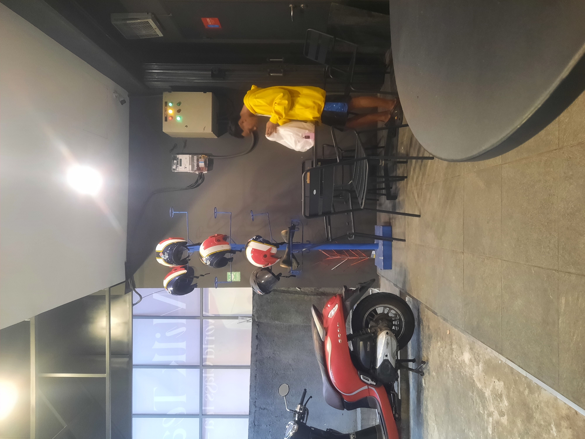 The image depicts an indoor scene, likely a cafe or waiting area, with an Asian person wearing a yellow hoodie and blue shorts standing near a table and chairs, holding a white plastic bag. In the background, several helmets are displayed on a rack. A red scooter is parked to the left of the scene. The environment appears to be a modern establishment with concrete walls and tiled flooring. The lighting suggests an indoor setting, possibly with artificial illumination. This scene is located in North Jakarta, Indonesia.