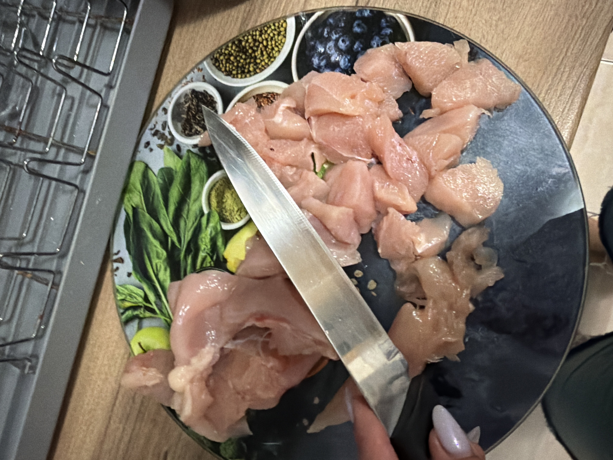 The image shows a close-up view of a person's hands preparing raw chicken. A knife is positioned over a plate with chopped chicken pieces. The plate has a decorative pattern featuring images of spinach, green apples, and various spices in small bowls, as well as blueberries. The background suggests a kitchen environment with a wooden countertop and a metal rack. The location is specified as Budva, Montenegro.