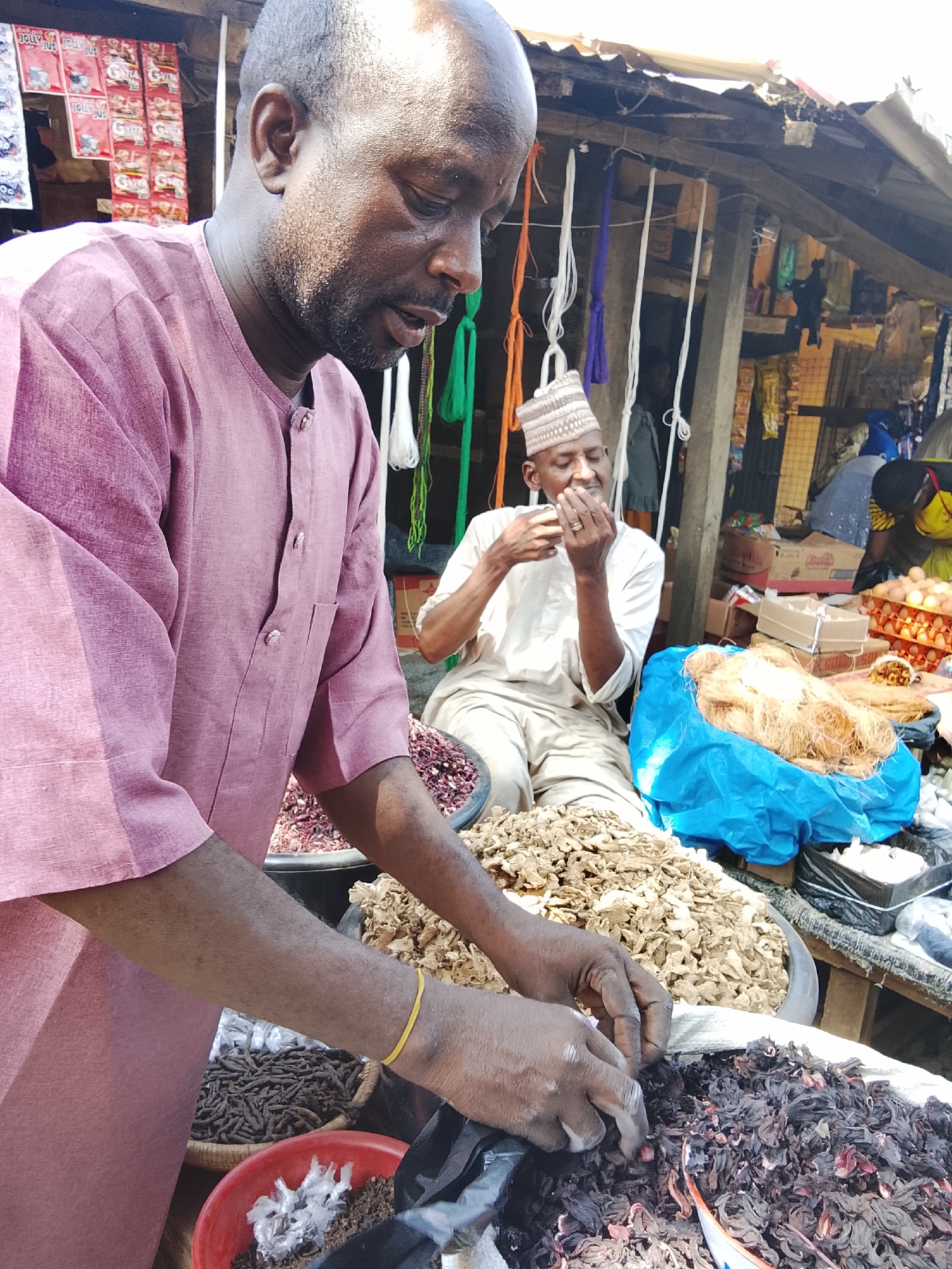 This image depicts an active market stall in Akwanga, Nigeria, under bright daylight. In the foreground, a bald, dark-skinned man with a short beard, wearing a long-sleeved pink/maroon tunic, stands behind a display of dried goods. His hands, with a yellow elastic band on the left wrist, are positioned over a large quantity of dark red, dried hibiscus flowers, appearing to sort or bag them. Directly to his right, a large black basin is filled with numerous light brown, dried, gnarled root pieces, consistent with ginger. Further right, a red plastic bowl contains white plastic shreds and dark granular material, and another woven basket holds a pile of dark, elongated dried items.
In the mid-ground, seated behind the stall, is a light-skinned man wearing a white tunic and a patterned white kufi cap. He holds a small, light-colored object to his mouth with both hands, seemingly consuming it. Behind him, the market structure consists of wooden beams supporting a corrugated metal roof. Various items are visible in the background, including red packets labeled "JOLLY JUS" hanging from a display, multiple colored cords (white, purple, green, orange) hanging vertically, cardboard boxes, and a tray of brown eggs. A large blue plastic bag contains a pile of light-colored, string-like material on the stall. Indistinct figures are visible deeper in the background.