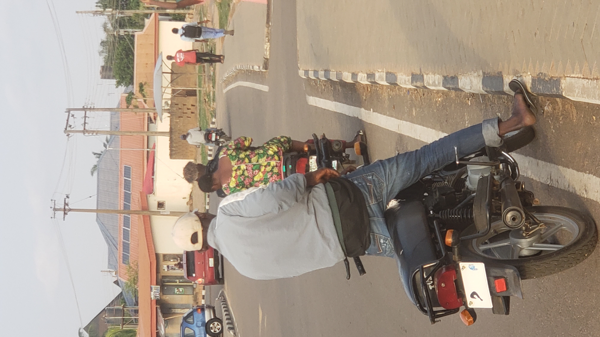 The image shows a street scene in Narayi, Nigeria, featuring two motorcycles carrying passengers. The foreground motorcycle has a rider wearing a white helmet, light blue jacket, blue jeans, and black shoes. A female passenger with a floral dress is seated behind the rider. The rear of the motorcycle is visible, including the license plate and taillight.
In the background, another motorcycle is visible, also with a rider and a passenger. Farther down the road, two individuals are walking away from the camera. Buildings with solar panels on the roofs and utility poles are visible on the left side of the street. Cars are parked along the roadside. The street is paved with white lane markings and a textured edge.