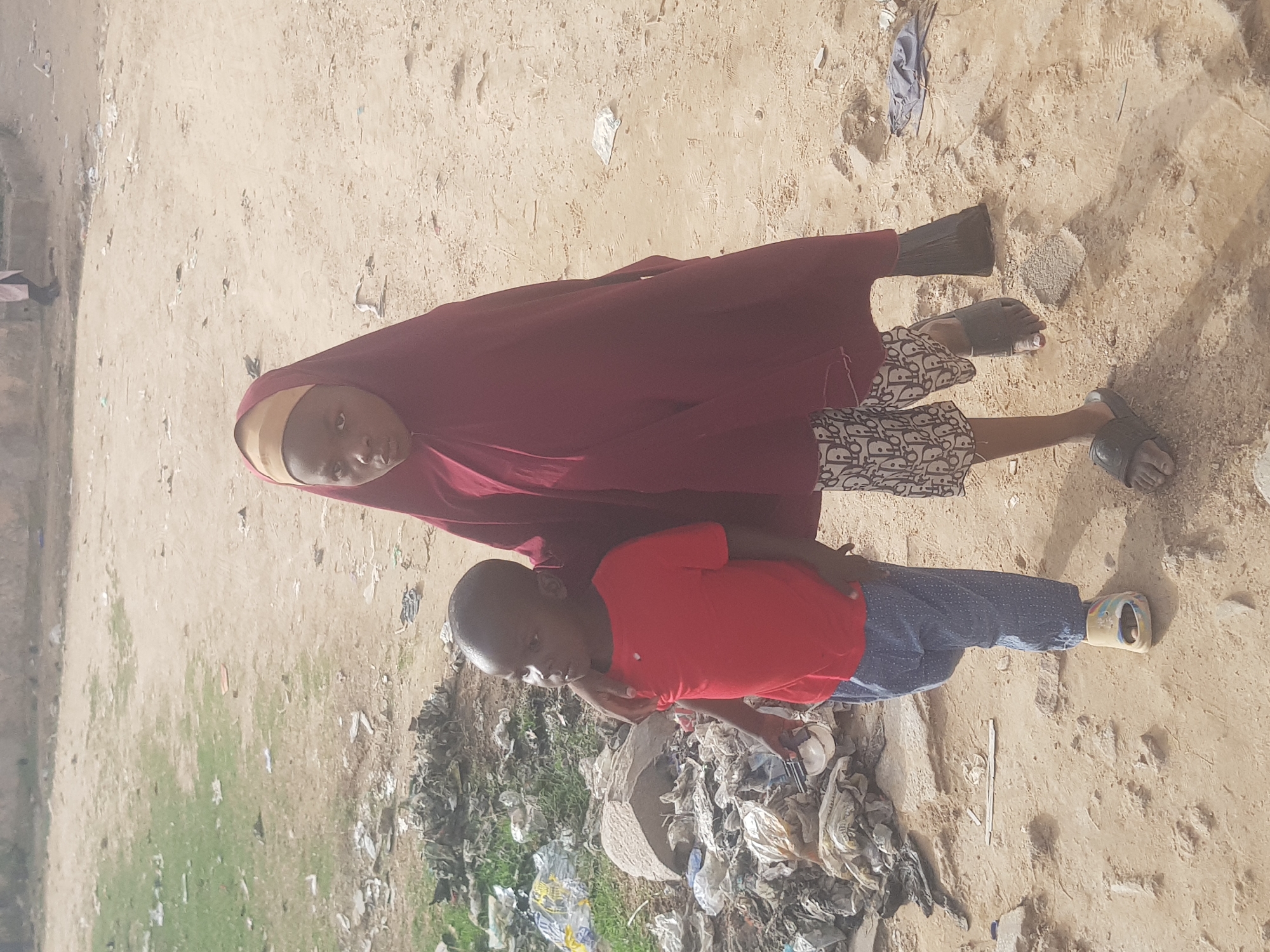 Two children are standing in a sandy area with visible debris and trash, possibly located in Dabawa, Nigeria. The older child is wearing a maroon hijab and a dress with a black and white pattern. The younger child is wearing a red shirt and blue pants. Both children are wearing sandals. The ground around them is sandy with scattered trash and some patches of green vegetation.