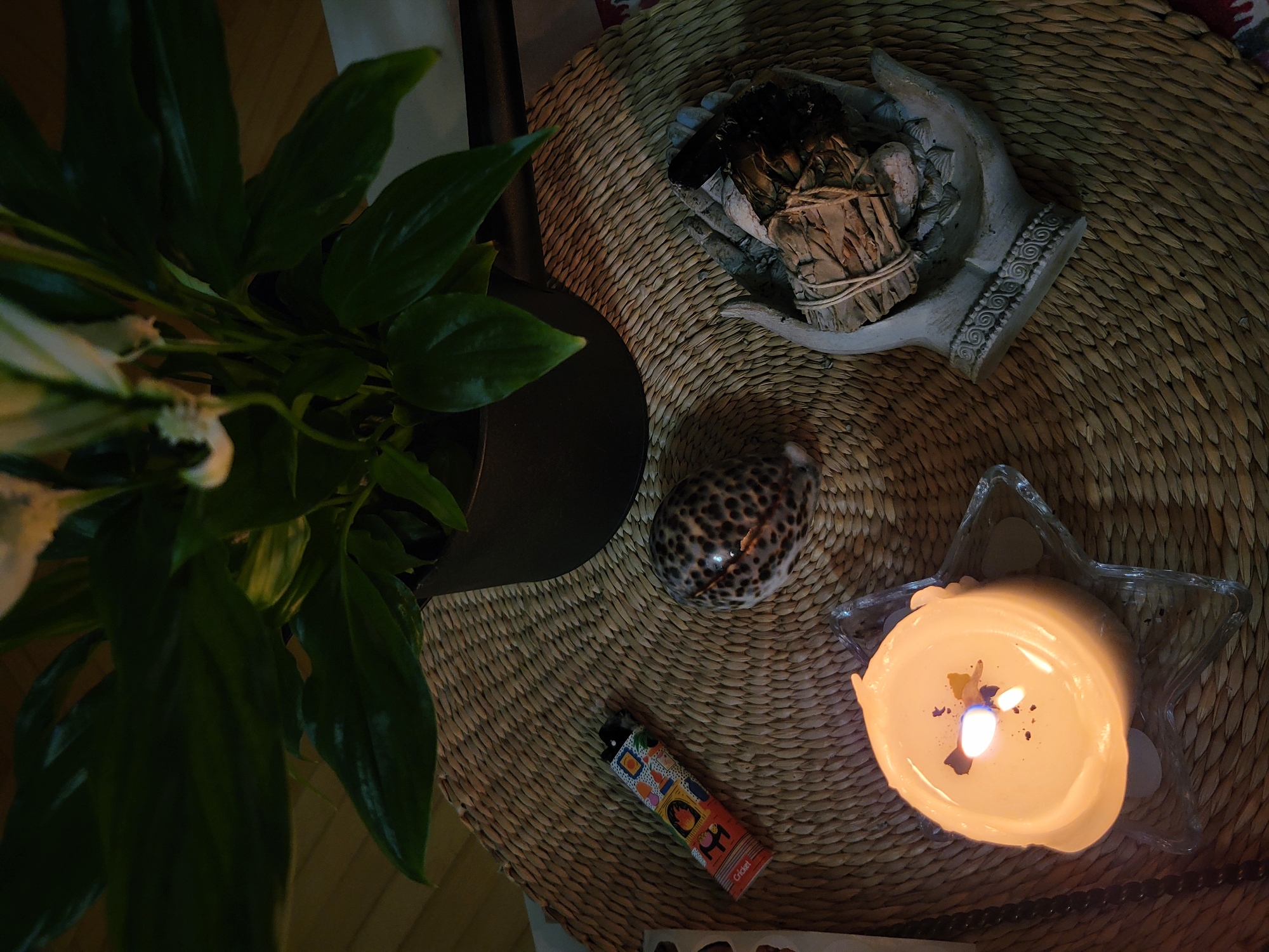 The image presents a high-angle view of a section of a woven mat, presumably a tabletop or decorative surface. On this mat are several objects: a potted peace lily plant with large green leaves to the left, a decorative set of hands holding a bundle of what appears to be dried sage and other elements, a shell with a leopard-like pattern, a colorful Cricket lighter, and a lit candle in a star-shaped glass holder. The lighting suggests an indoor setting, with the candle providing some illumination. The scene seems to convey a spiritual, new age or meditative atmosphere. There is no information to suggest the image was taken in Budva, Montenegro.