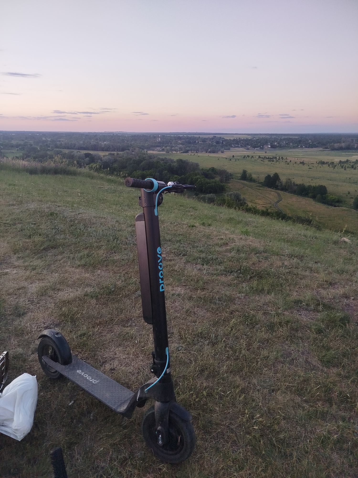 An electric scooter with the brand name "proove" in blue lettering is parked on a grassy hill. The scooter is black with blue accents and has a vertical battery pack. In the background, a wide valley unfolds with a village nestled among trees, and a vast expanse of fields stretching towards the horizon. The sky is a soft gradient of pink and purple, indicating either dawn or dusk. A white plastic bag is visible to the left of the scooter. The scene is set in Ustyvytsia, Ukraine.