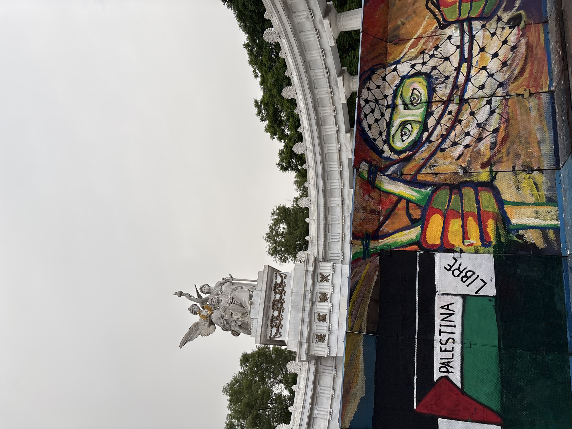 The image captures a section of the Monumento a la Raza in Mexico City, Mexico. In the foreground, a mural is painted on a protective barrier. The mural features the Palestinian flag on the left, with the words "PALESTINA LIBRE" written across it in black capital letters. To the right of the flag, the mural depicts a person wearing a keffiyeh, with their eyes prominently visible. The background of the mural is a warm orange and brown hue, with other colorful abstract elements. Behind the mural and barrier, a white marble monument with classical architectural elements is visible. This monument features sculptures of figures, including one with wings. The sky above is overcast and pale. Trees are visible behind the monument.
