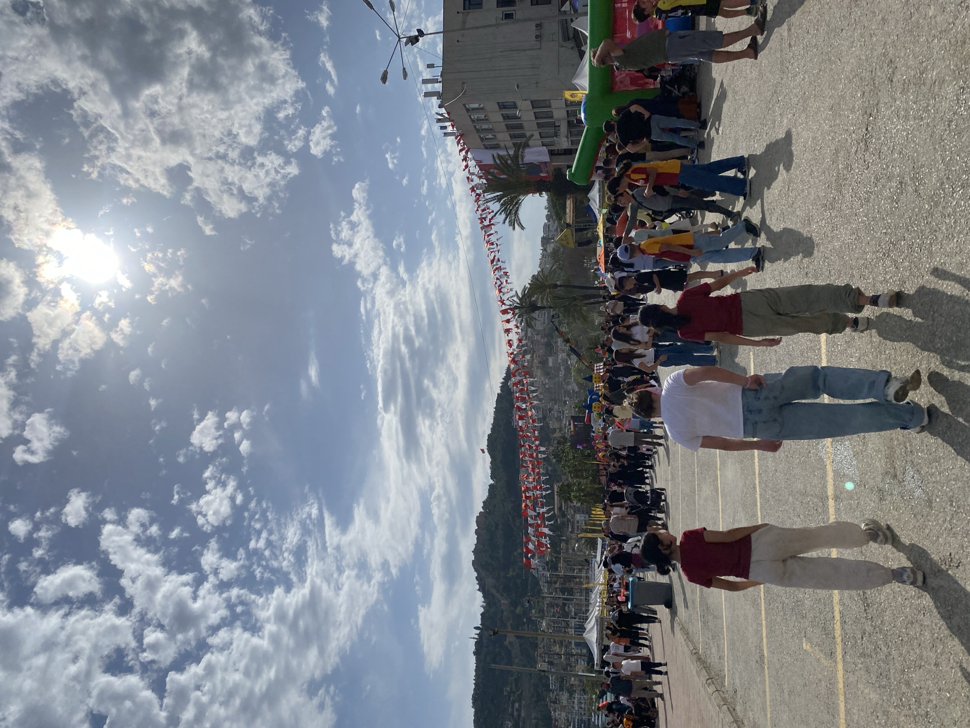 The image captures a scene in Alanya, Turkey. The sky is partially covered with clouds, with the sun shining through. A crowd of people is gathered along a paved area marked with yellow lines. Some individuals are walking. Buildings and palm trees line the background. Turkish flags are strung overhead. A mountain rises in the distance, densely populated with buildings. An inflatable structure is present.